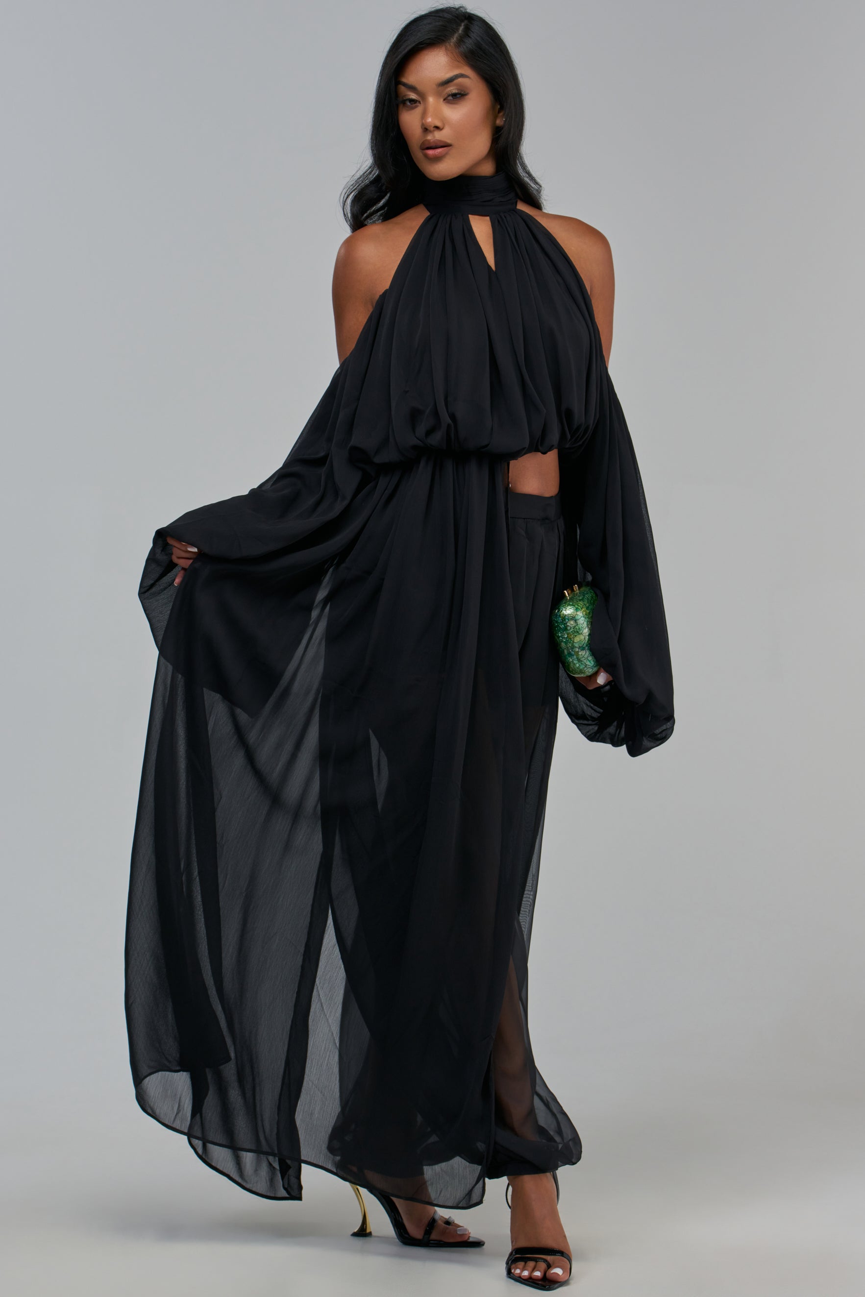A person wears the LEAN ON ME CHIFFON BLOUSE IN BLACK, featuring a cold shoulder design in sheer chiffon with a high, cutout neckline and long, voluminous sleeves, paired with matching wide-leg trousers and black heeled sandals.