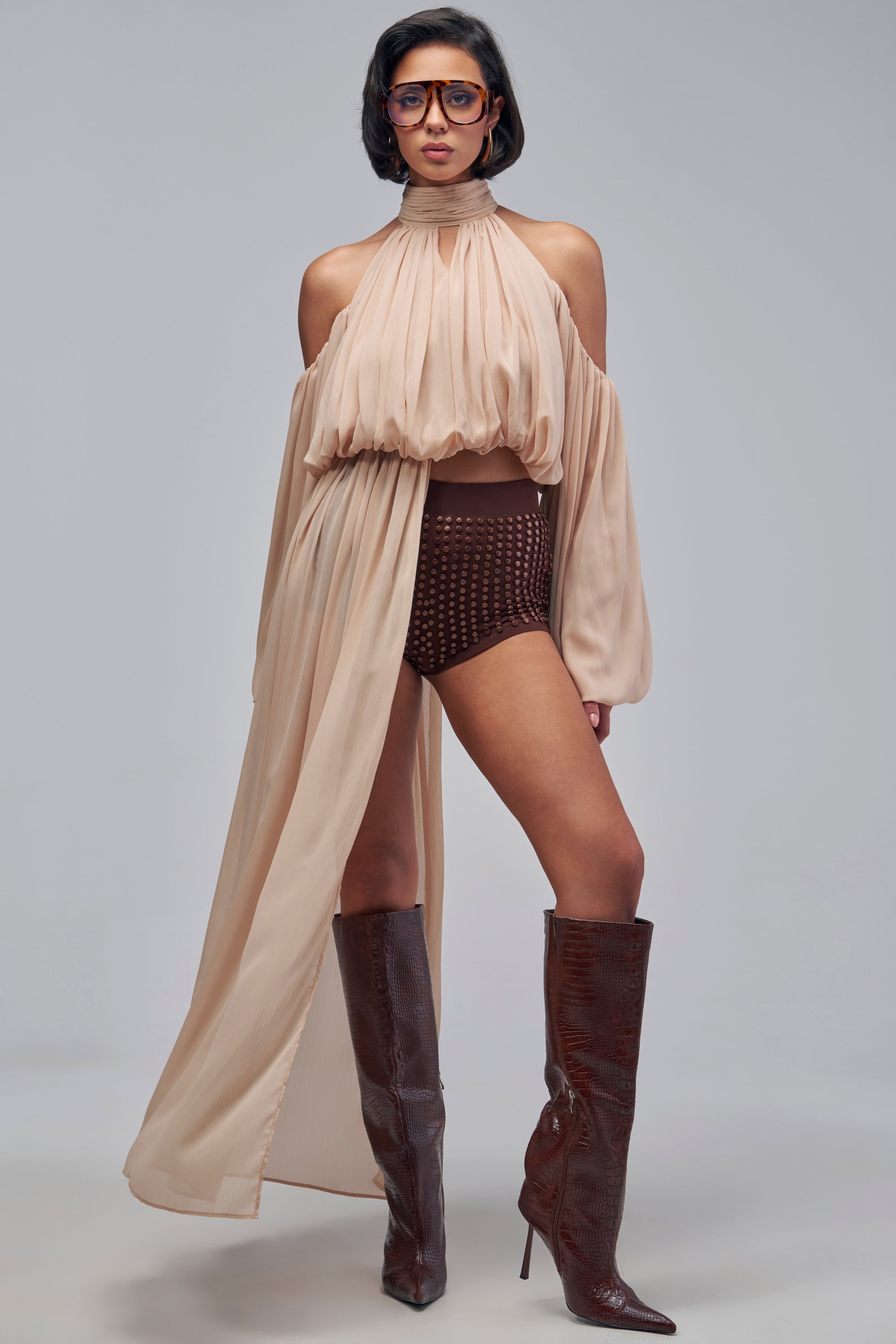 A person wearing a beige sheer chiffon LEAN ON ME CHIFFON BLOUSE with a cold shoulder design and long skirt overlay, paired with brown high-waisted shorts and knee-high brown boots.