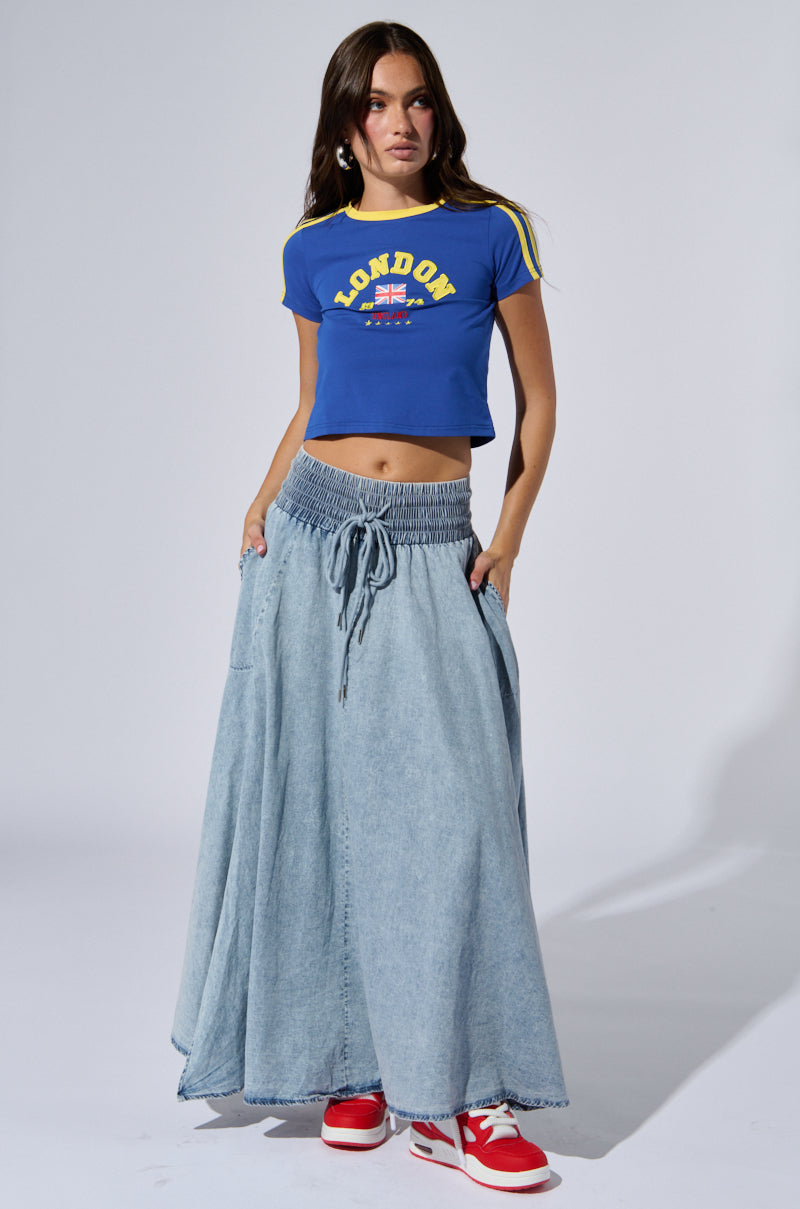 A woman stands against a plain background, wearing a blue crop top with "LONDON" and an English flag, the Alex acid wash denim maxi skirt with its high-rise elastic waistband, and red and white sneakers. She has long straight hair and her hands are in her pockets.