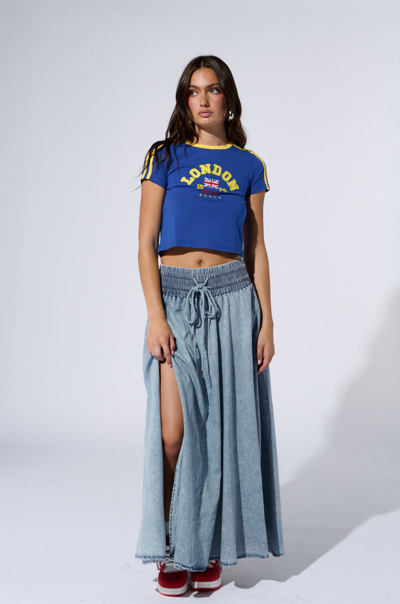 A young woman stands facing forward against a plain background. She wears a blue crop top with yellow stripes and "London" written on it, paired with the ALEX ACID WASH DENIM MAXI SKIRT featuring a high side slit. She has long brown hair and red shoes.