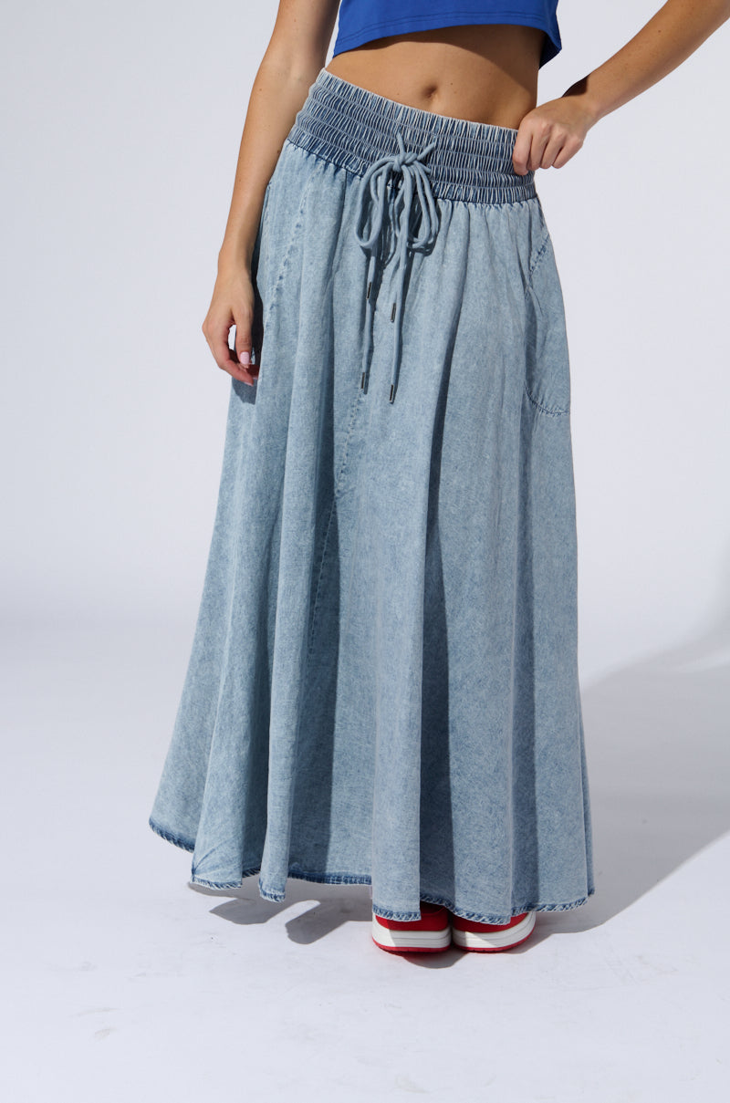 A person wearing the ALEX ACID WASH DENIM MAXI SKIRT with a high-rise elastic waistband and sneakers. The top portion of the person is mostly cropped out of the image, showing only their midsection and legs. The background is a plain, light gray.