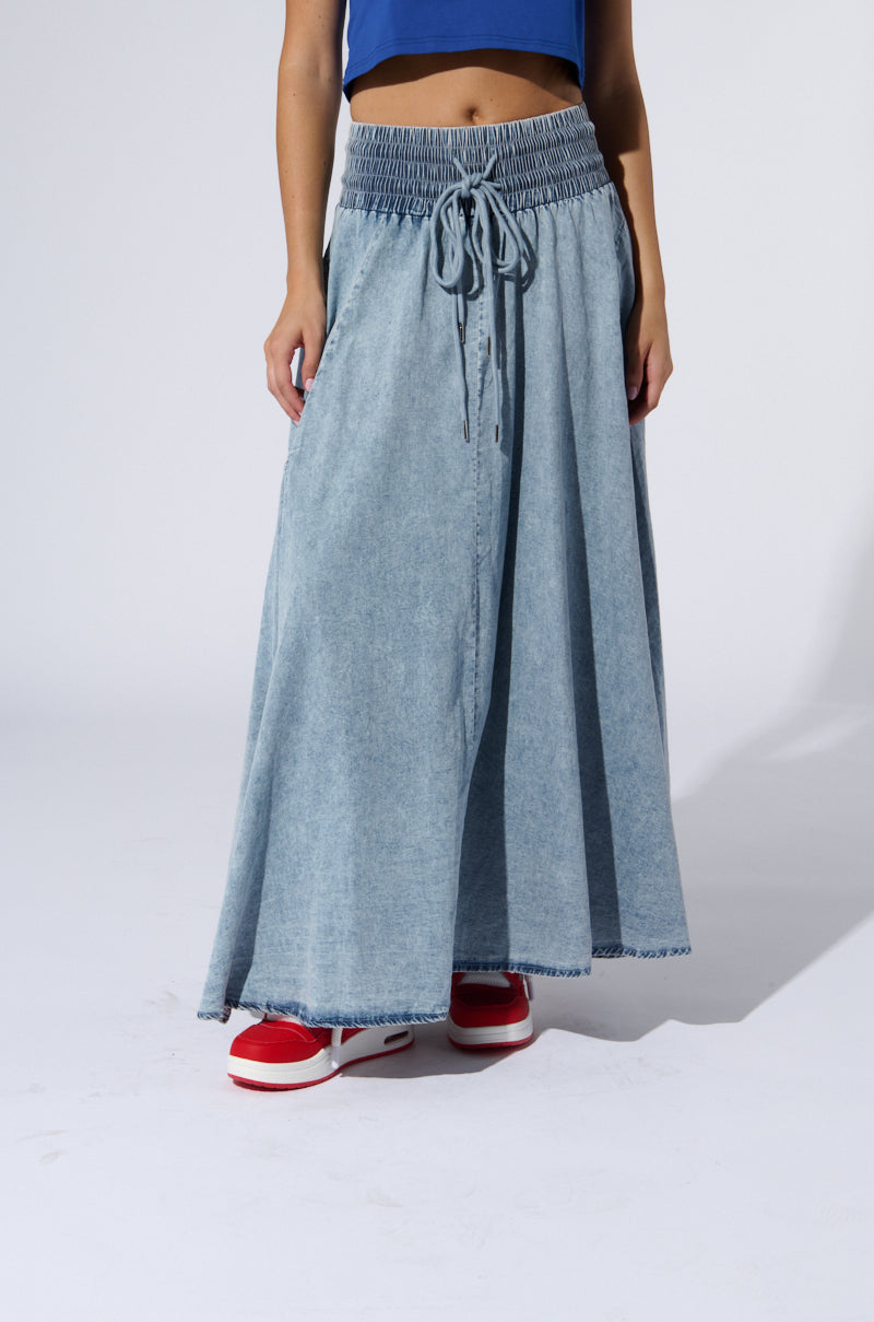 A person is standing against a plain background wearing the ALEX ACID WASH DENIM MAXI SKIRT. The person also wears a cropped, sleeveless top and red sneakers with white soles. The face is not visible in the image.