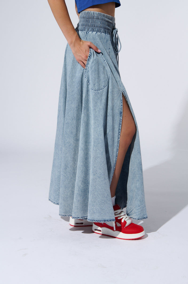 A person wearing the ALEX ACID WASH DENIM MAXI SKIRT, featuring side slits and a high-rise elastic waistband. They have one hand in their pocket and are also sporting red and white sneakers. The background is plain white.