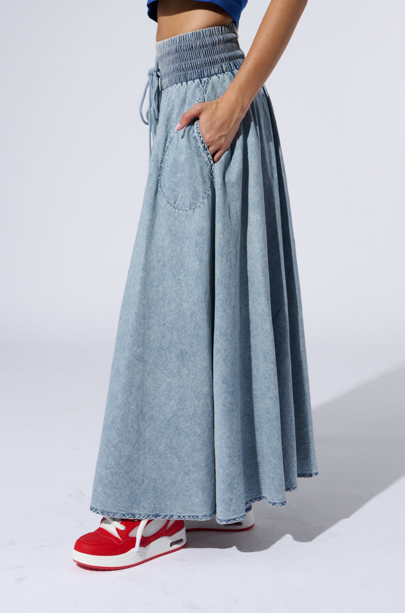A woman wearing the ALEX ACID WASH DENIM MAXI SKIRT, which features a high-rise elastic waistband and pockets, along with red sneakers stands against a plain background. Only the lower part of her torso and legs are visible in the image.