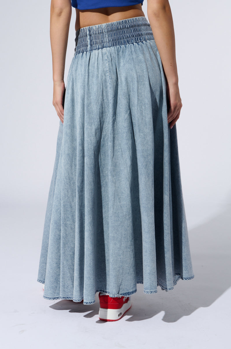 A person is standing with their back to the camera, dressed in the ALEX ACID WASH DENIM MAXI SKIRT, which showcases a high-rise elastic waistband. The skirt is long, flares out towards the bottom, and features a stylish side slit. Their look is completed with red and white sneakers against a plain, light-colored background.