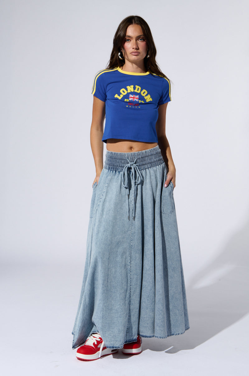 A woman with long brown hair stands against a plain background, wearing a blue crop top with "London" written on it, an ALEX ACID WASH DENIM MAXI SKIRT featuring a drawstring waist and side slit, and red sneakers. Her left hand is in her pocket as she gazes directly at the camera.