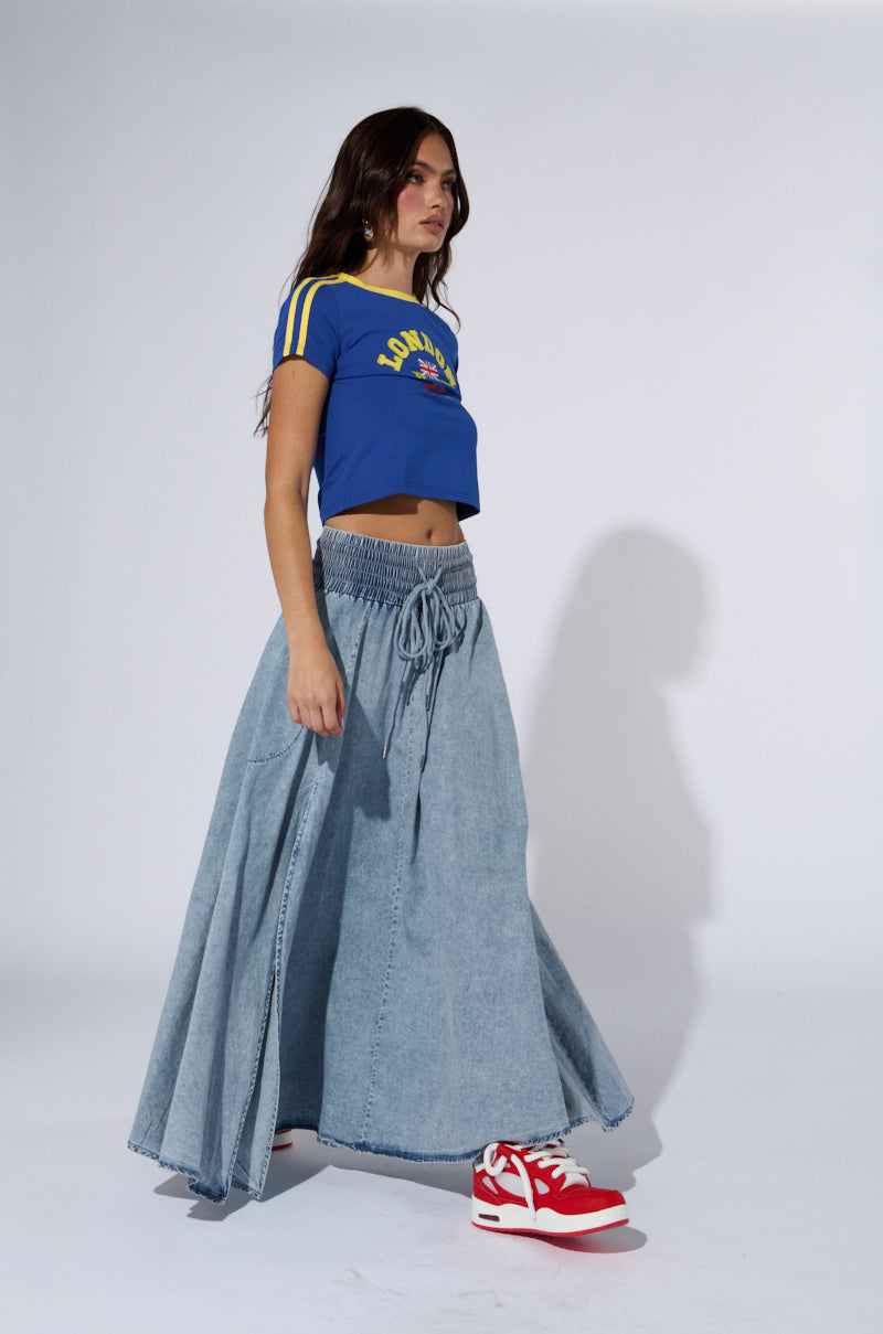 A woman is wearing a cropped blue T-shirt with yellow stripes on the sleeves and a graphic design, paired with the ALEX ACID WASH DENIM MAXI SKIRT, which features a side slit. She has red and white sneakers on her feet. She walks and looks to her left against a plain white background.