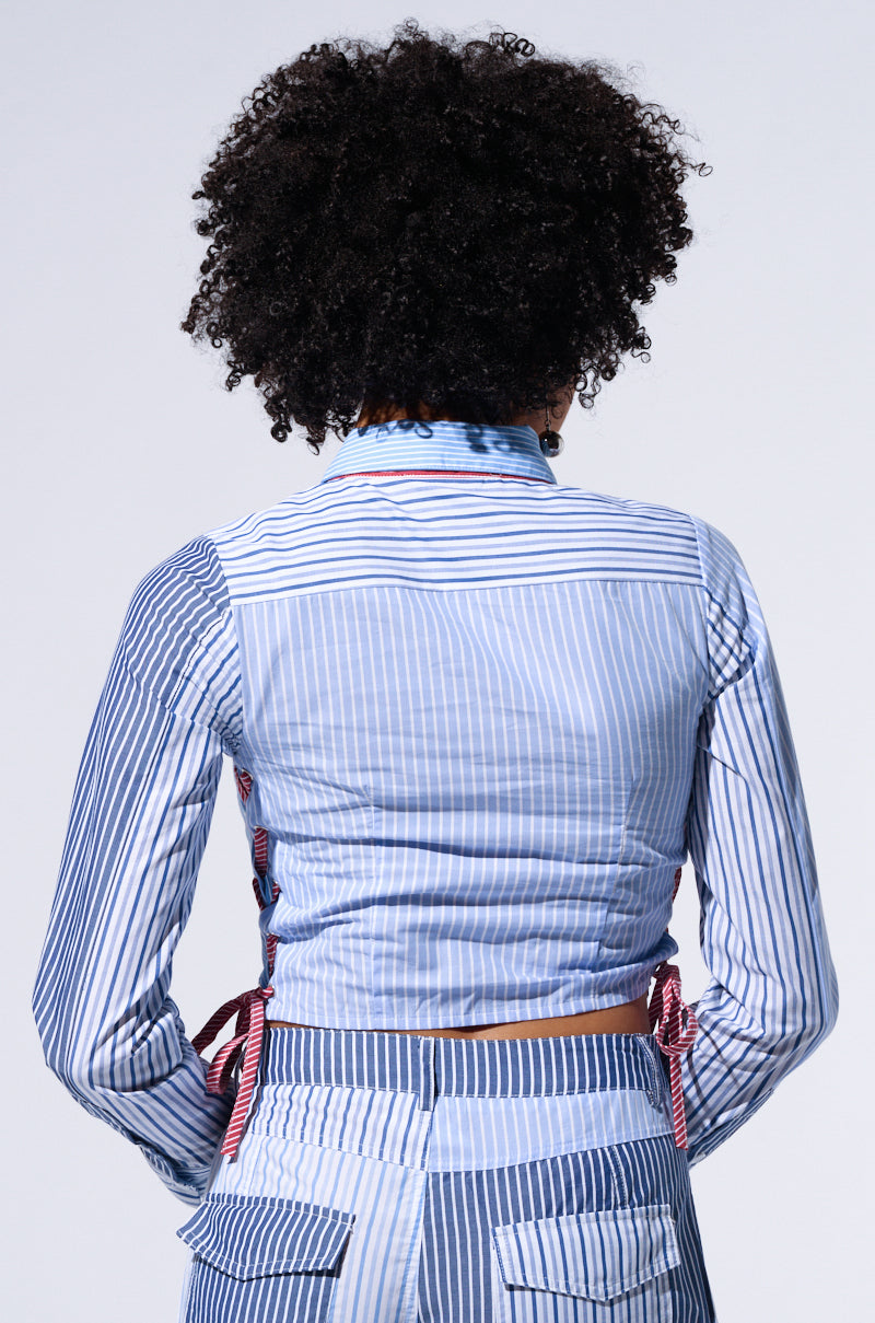 A person with curly hair is seen from behind, wearing the BUSINESS GIRL CROPPED BUTTON DOWN BLOUSE in a light blue and white striped pattern with red side ties. The blouse is paired with matching high-waisted pants featuring patchwork stripe detailing and patch pockets.