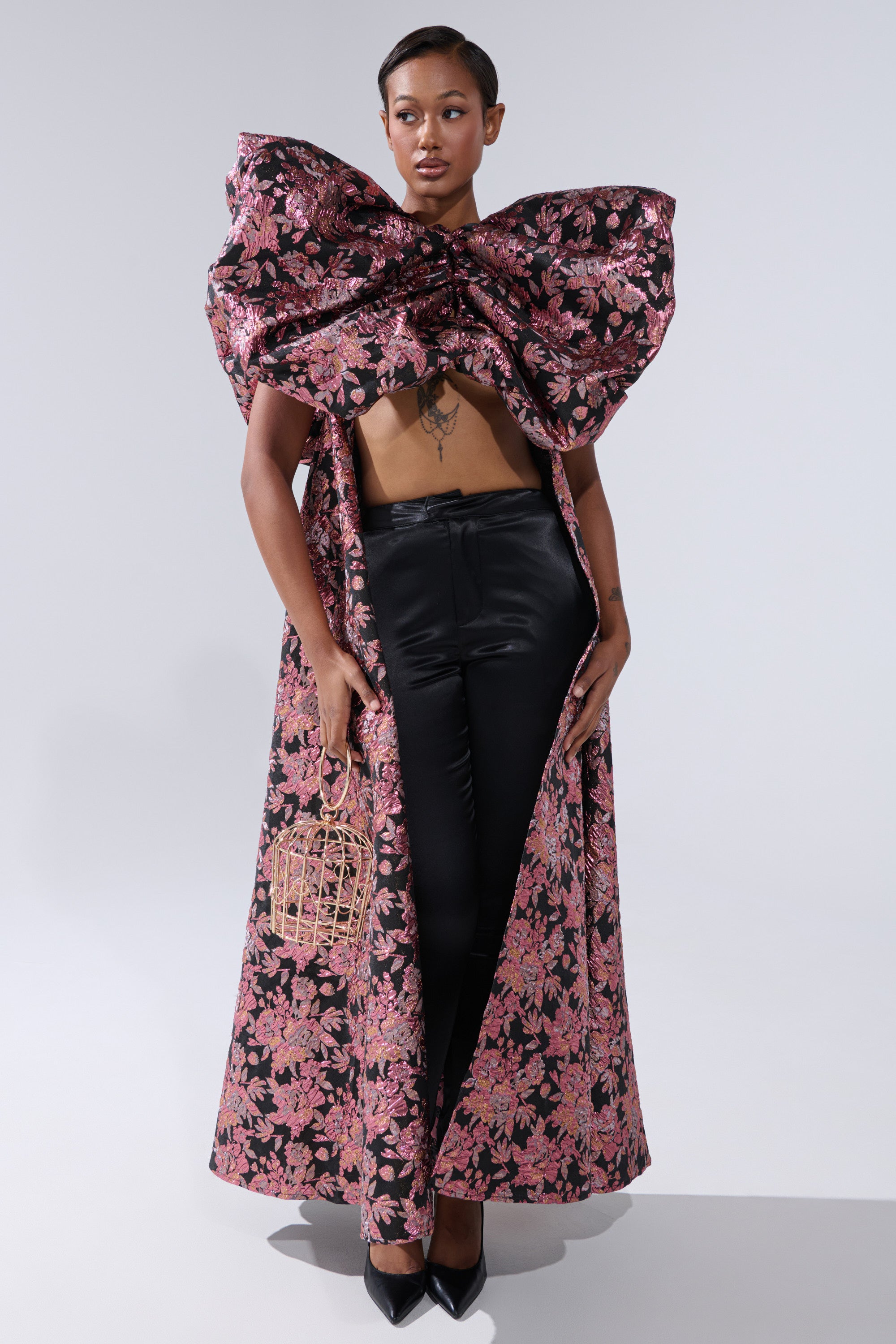 A woman exudes confidence in black pants and the DIVINE INTERVENTION BROCADE TOP, styled as a floral cape with an oversized bow that reveals her midriff. She accessorizes with a cage-shaped handbag and pointed black shoes. 0