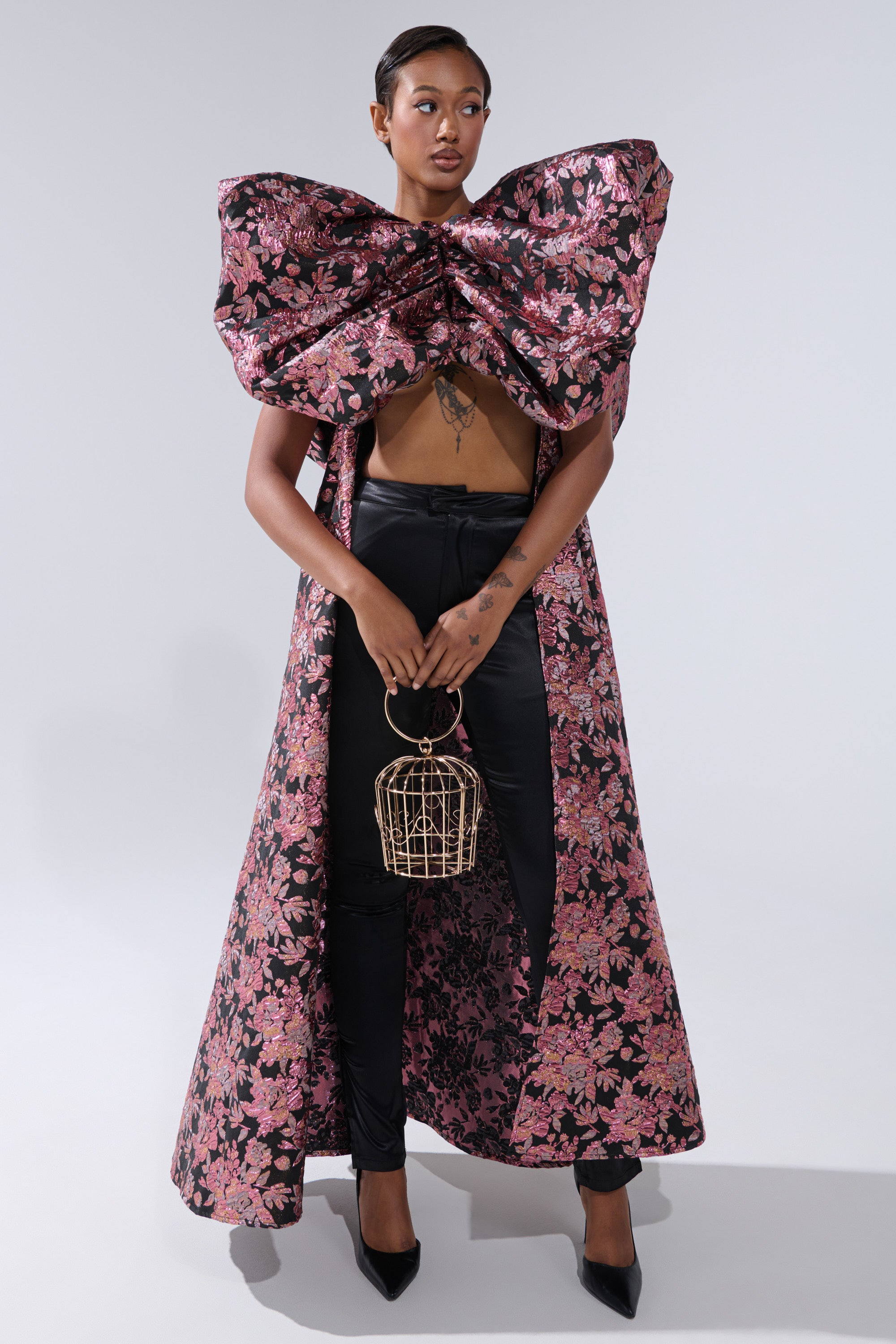 A woman wears the DIVINE INTERVENTION BROCADE TOP—featuring dramatic pink and black florals, puffed shoulders, and a large matching bow—paired with black pants, heels, and a birdcage-shaped purse against a light gray background. 1
