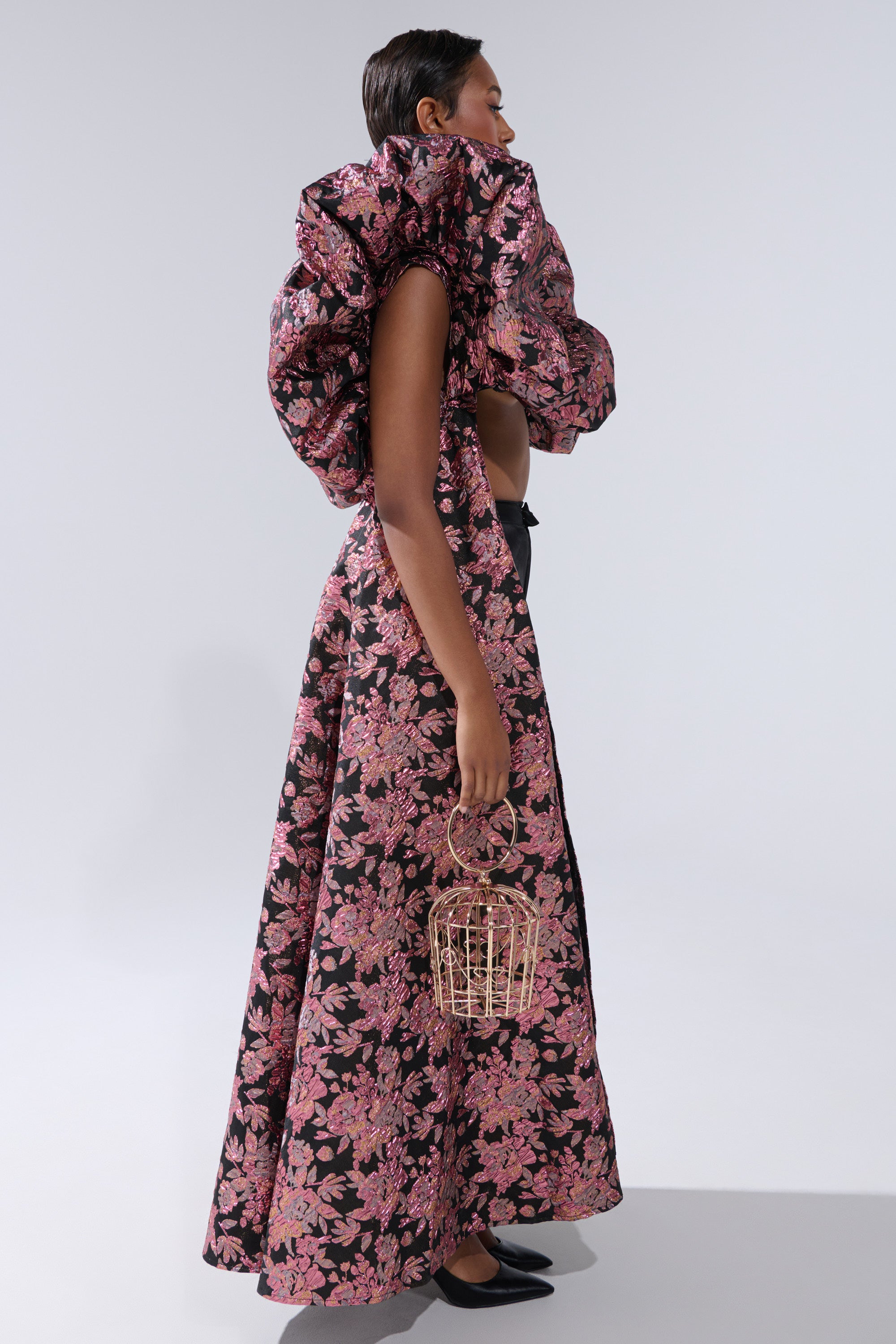 A woman in the DIVINE INTERVENTION BROCADE TOP, a dramatic black and pink floral piece with puffed shoulders and an oversized ruffled collar, stands in profile holding a small cage-like handbag, wearing black shoes against a light gray background. 3