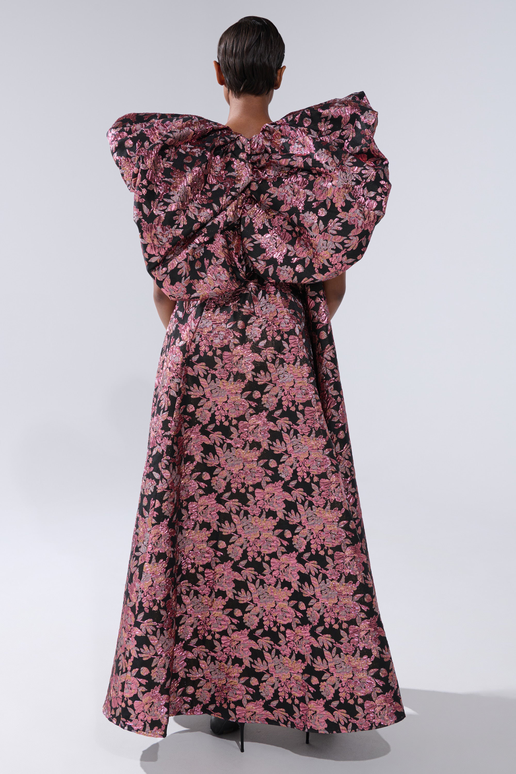A person stands facing away, wearing the DIVINE INTERVENTION BROCADE TOP with dramatic, oversized puffed shoulders and a floor-length black dress featuring a pink floral pattern. The background is plain and light-colored. 4