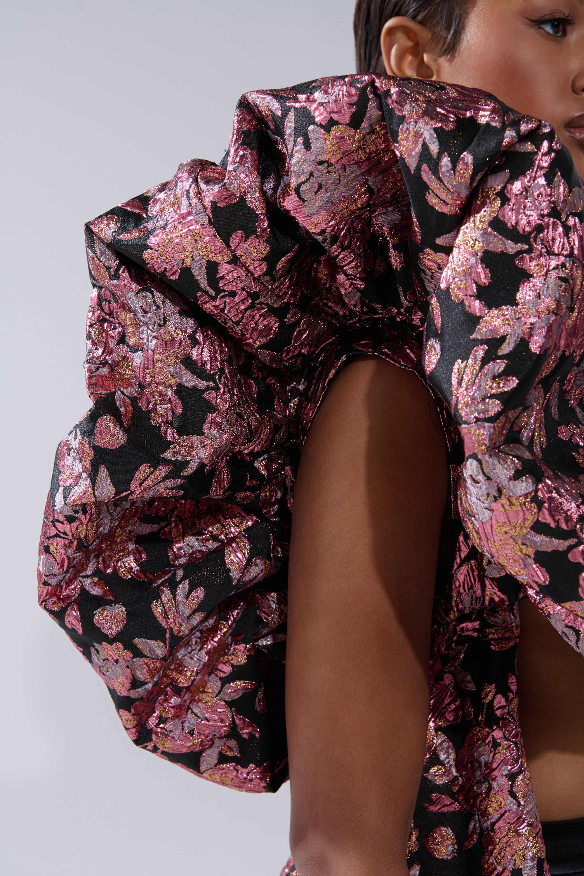 A person partially visible in the DIVINE INTERVENTION BROCADE TOP, featuring dramatic black and pink metallic floral ruffles, with only their face, shoulder, and the textured blouse shown against a neutral background. 5