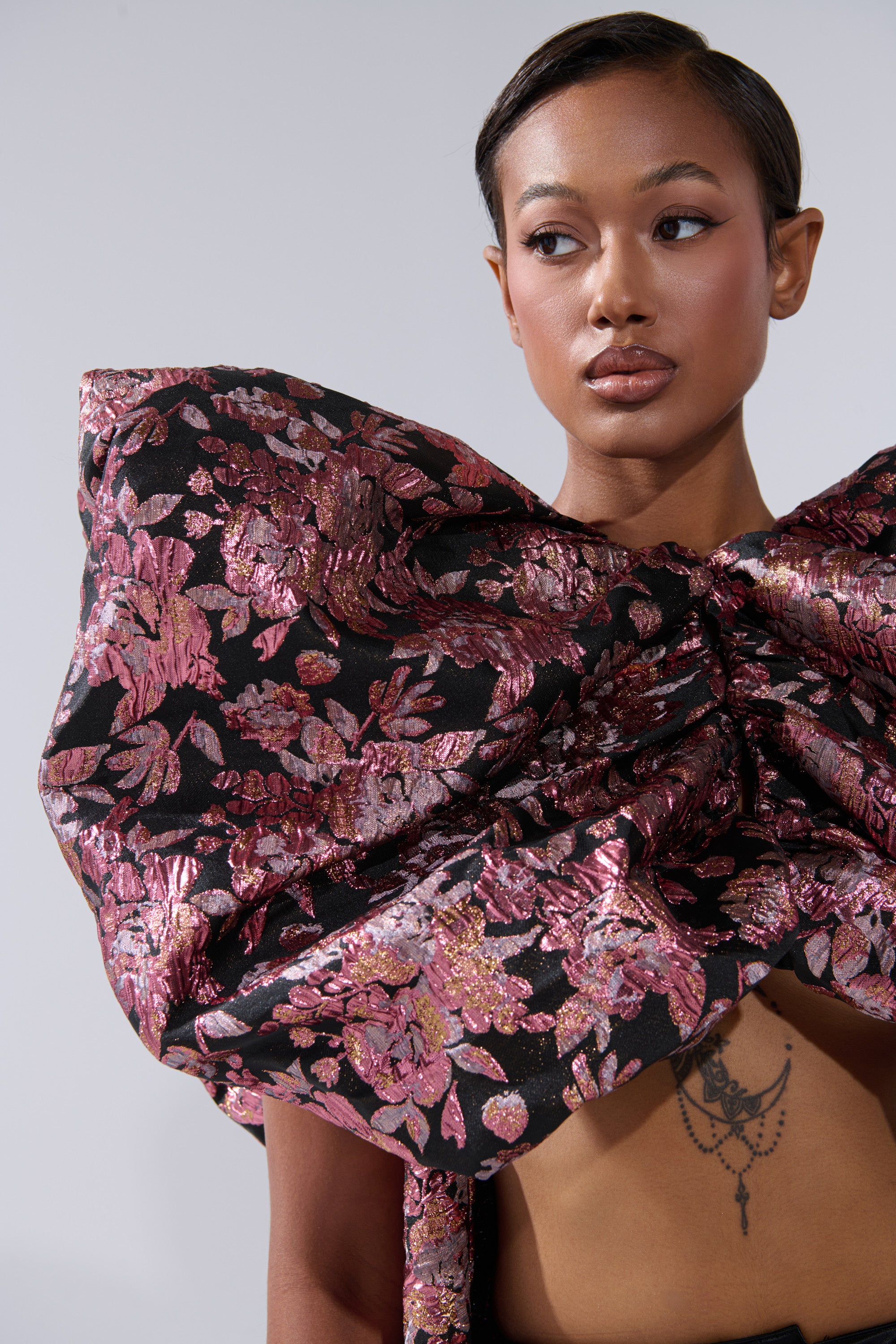 A woman with sleek hair looks to the side, wearing an oversized metallic pink DIVINE INTERVENTION BROCADE TOP with a floral finish that reveals her midriff and chest tattoo. The background is plain light gray. 6