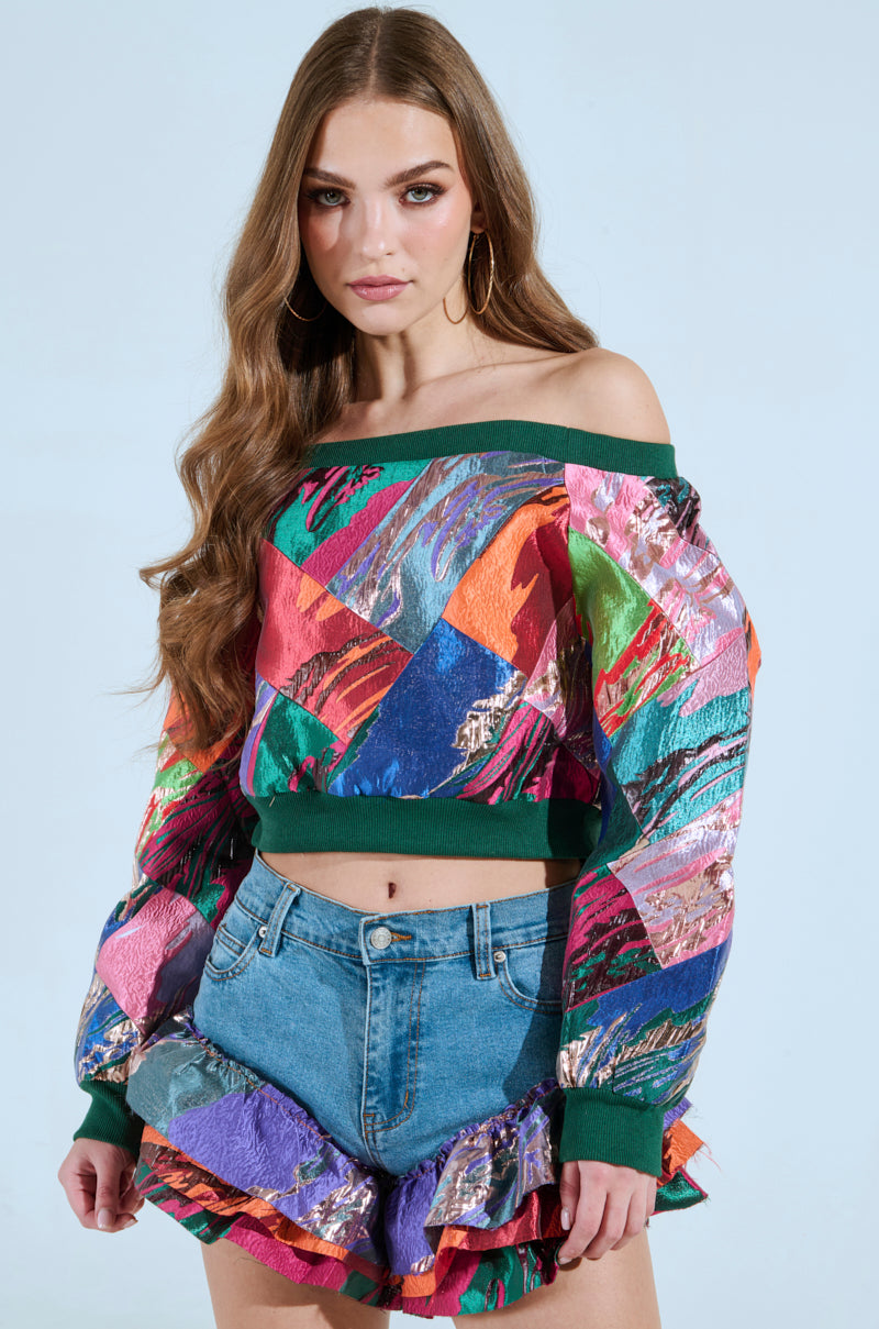 A woman with long brown hair wears the LIFE IS BUT A DREAM OFF THE SHOULDER MULTI PRINT SWEATSHIRT with metallic elements and technicolor brocade, paired with denim shorts featuring a ruffled hem, set against a light blue background.