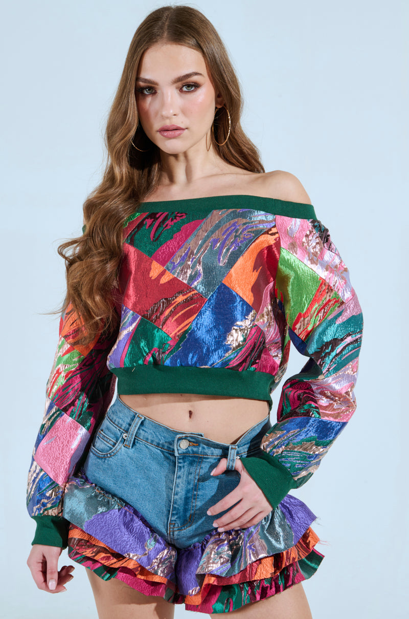A woman with long wavy hair wears the "Life Is But A Dream Off the Shoulder Multi Print Sweatshirt" and denim shorts with ruffled, multicolored edges, set against a light blue background. Hoop earrings complement her confident expression.
