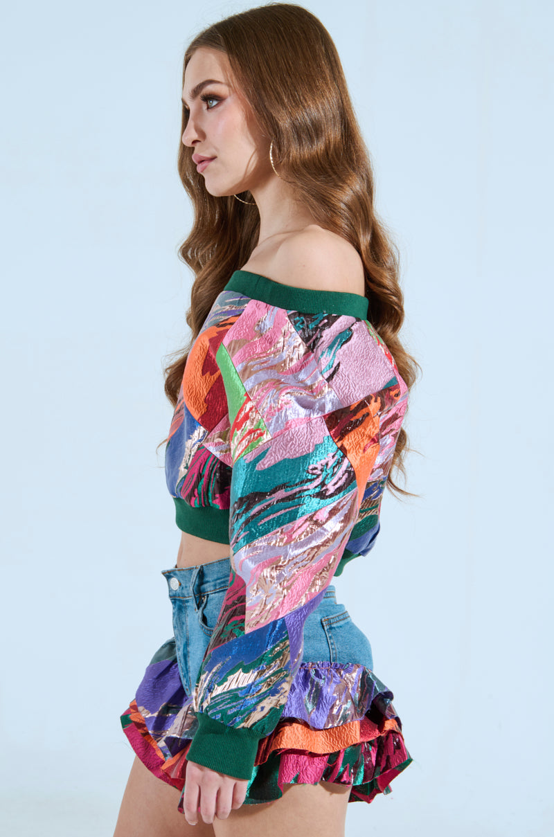 A woman with long brown hair poses against a light blue background, wearing the LIFE IS BUT A DREAM OFF THE SHOULDER MULTI PRINT SWEATSHIRT and ruffled denim shorts. Her expression is calm as she looks to her left.