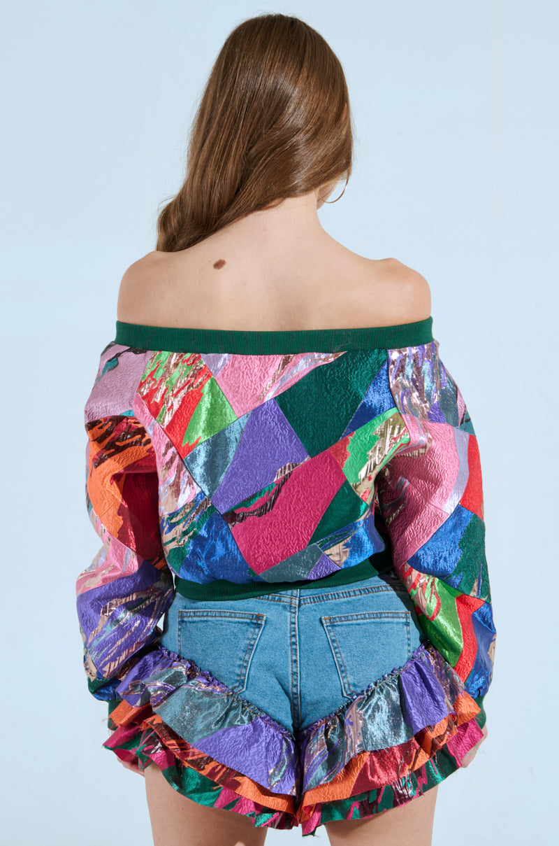 A person faces away from the camera, donning a LIFE IS BUT A DREAM OFF THE SHOULDER MULTI PRINT SWEATSHIRT and denim shorts with multicolored ruffles, set against a plain light blue background.