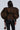 A person with long dark hair is seen from the back, highlighting the V-neckline of their "Blow Him A Kiss Belted Cheetah Blouse" featuring a brown and black cheetah print with large puffed sleeves. They carry a small matching bag and wear black leather pants, all set against a neutral backdrop that accentuates their chic ensemble.