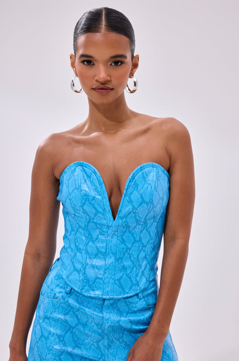 A woman wears the ELECTRIC SNAKE CORSET, a strapless blue snakeskin top with a deep V neckline, paired with matching pants. Her sleek hair and large hoops complete the look as she poses against a light background.