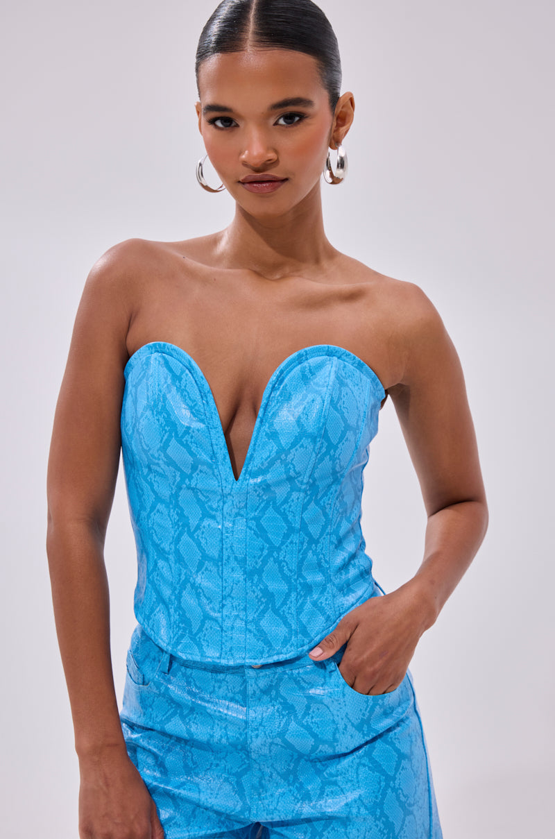 A woman with slicked-back hair wears large hoop earrings and the strapless blue ELECTRIC SNAKE CORSET with a deep V neckline, facing forward with one hand in her pocket against a plain backdrop.
