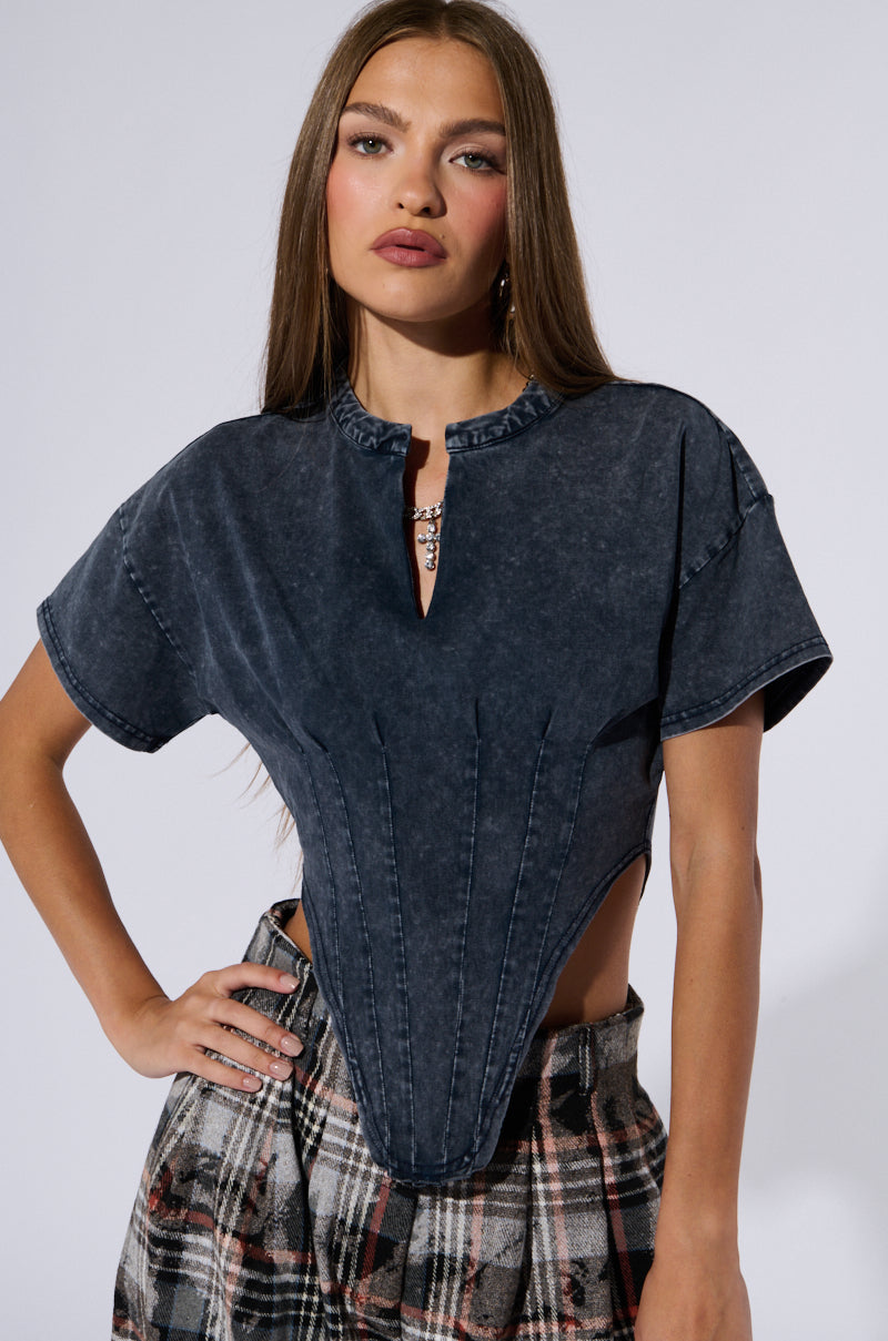 A woman with long brown hair is posing for the camera. She is wearing a SEIZE THE DAY SHORT SLEEVE CORSET T-SHIRT paired with plaid high-waisted pants. She has a neutral expression on her face.