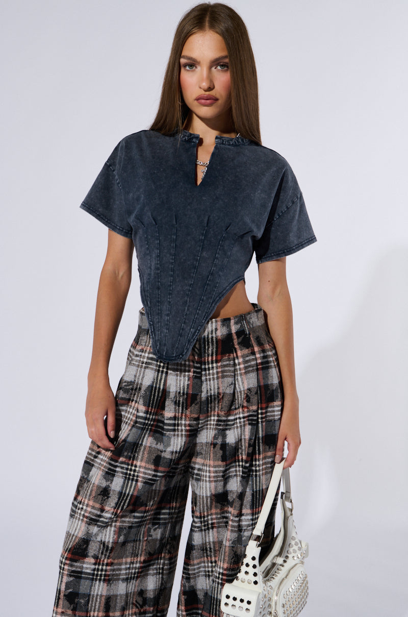 A woman with long brown hair is wearing a SEIZE THE DAY SHORT SLEEVE CORSET T-SHIRT in dark blue with a unique cut-out design, paired with high-waisted wide-leg plaid trousers. She is holding a white handbag and standing against a plain light-colored background.