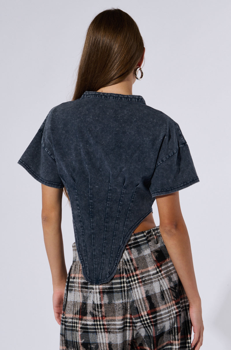 Rear view of a person with long brown hair wearing plaid pants and the SEIZE THE DAY SHORT SLEEVE CORSET T-SHIRT. The black denim top, inspired by a corset design, features a unique, structured back. The person is standing against a plain background.