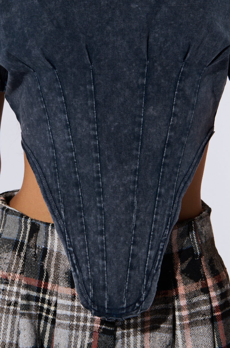 Close-up of a person wearing the "SEIZE THE DAY SHORT SLEEVE CORSET T-SHIRT," a dark distressed, mineral wash crop top with tailored stitching details that create a structured look, paired with plaid pants in shades of grey and black with hints of red.