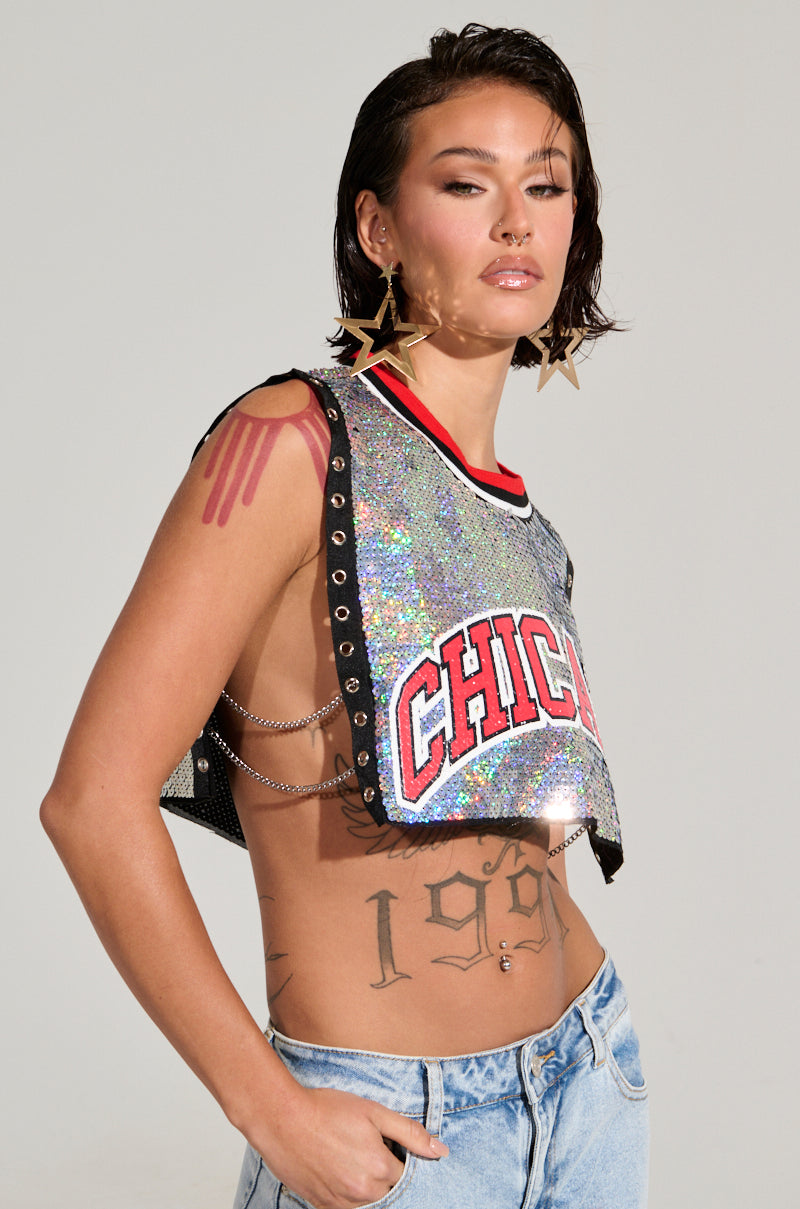 Sporting short hair, an individual is dressed in the CHICAGO SEQUIN CROPPED TANK—a captivating iridescent silver piece featuring chain details and open sides. Large star-shaped earrings, visible arm tattoos, and jeans add to their ensemble, while their confident expression contrasts with the plain backdrop.