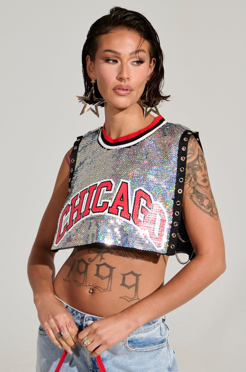 A person with short hair is adorned in large star-shaped earrings and a CHICAGO SEQUIN CROPPED TANK, shimmering with iridescent silver sequins. Displaying visible tattoos, they grasp red strings, complementing the ensemble with light blue jeans against a simple background, radiating effortless style.