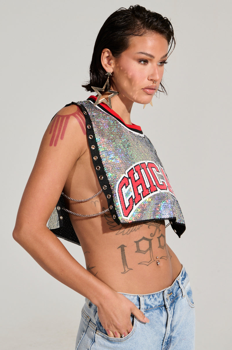 A person with short hair stands against a plain background, wearing the CHICAGO SEQUIN CROPPED TANK—an iridescent silver sequin cropped top with "CHICAGO" boldly printed across it—and denim jeans. They sport large star-shaped earrings, several tattoos on their arms and torso, with the tank's stylish open sides adding a hint of edge.