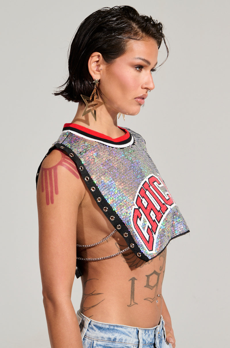 A person with short dark hair is wearing large star earrings and the CHICAGO SEQUIN CROPPED TANK, an iridescent silver sequin crop top emblazoned with "Chicago." Their tattooed arms and body are displayed through the open sides of this tank, which pairs perfectly with light blue denim jeans against a plain background.