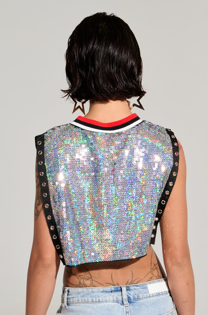 A person with short, dark hair is turned away from the camera, showcasing a CHICAGO SEQUIN CROPPED TANK adorned with iridescent silver sequins, black trim, and grommet details. They are accessorized with star-shaped earrings and paired with light blue jeans.