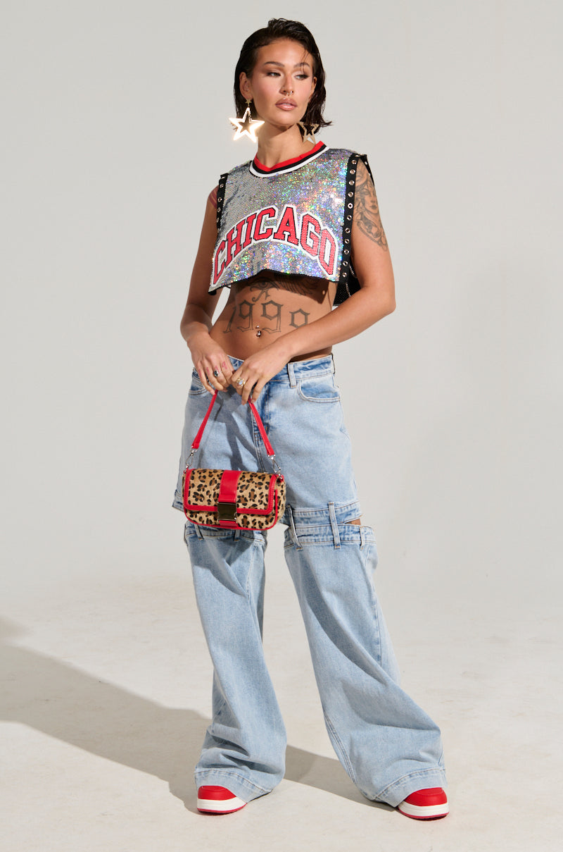 A person stands confidently in the CHICAGO SEQUIN CROPPED TANK, shimmery with iridescent silver sequins and featuring open sides. It is paired with wide-leg jeans and red sneakers. They accessorize with a leopard-print handbag and star-shaped earrings, while their midriff and arms are adorned with tattoos.