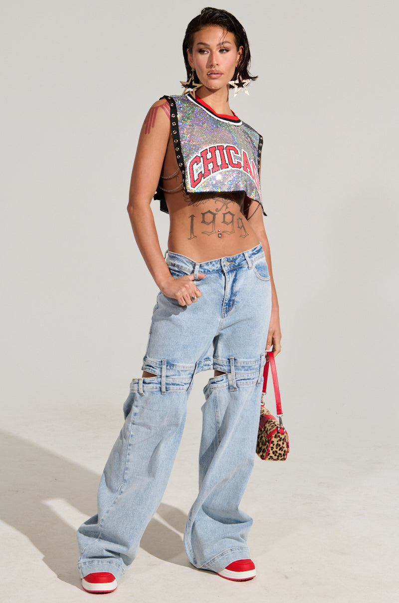 Wearing the CHICAGO SEQUIN CROPPED TANK embellished with shimmering iridescent silver sequins and a pair of distressed wide-leg jeans, a person confidently carries a leopard print bag. They step out in red shoes, showcasing their "1998" tattoo across their torso. Their look is completed with star earrings and a chic short hairstyle.