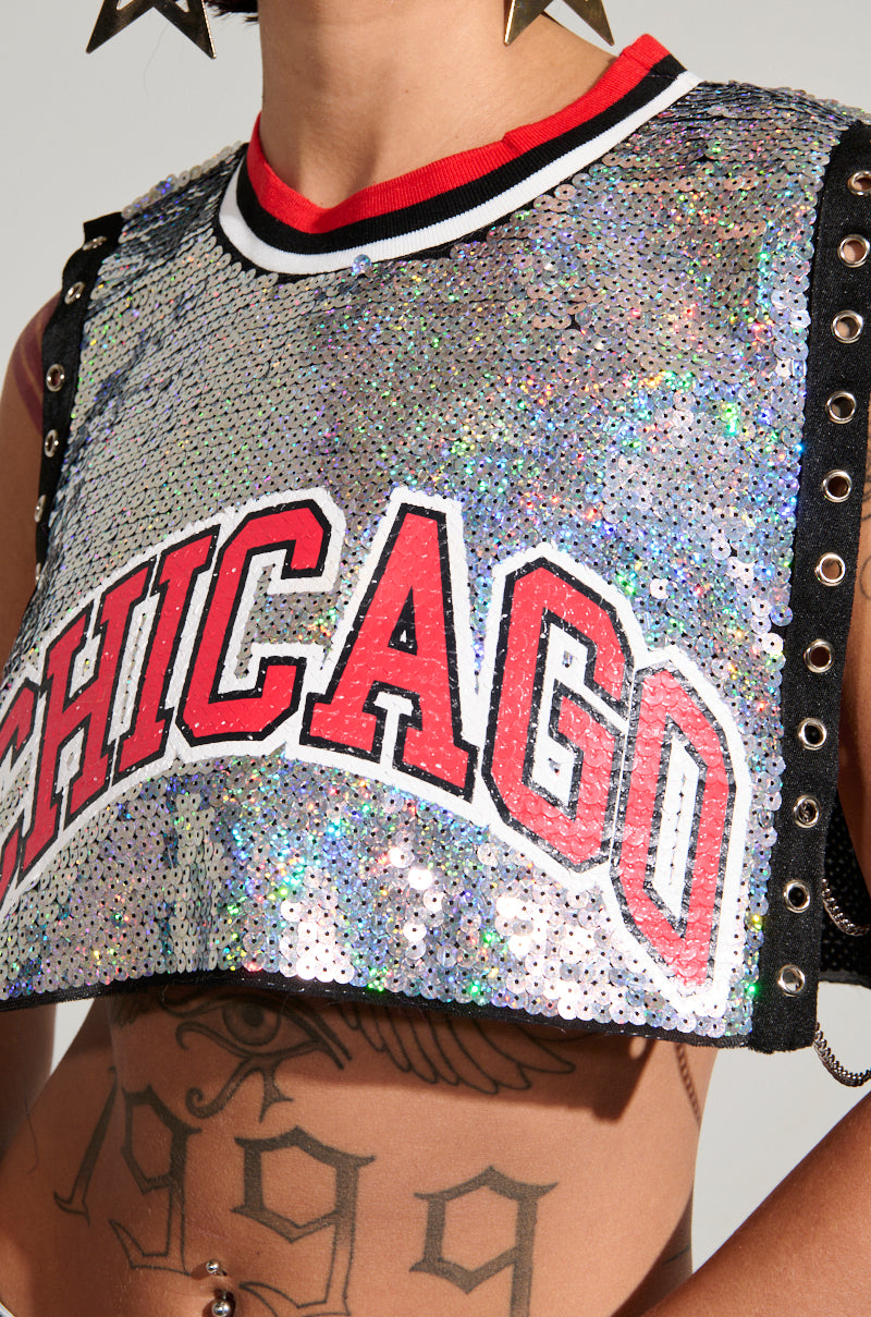 A person is dressed in the chic CHICAGO SEQUIN CROPPED TANK, which dazzles with iridescent silver sequins and displays the word "CHICAGO" in striking red letters. This top includes black side lacing and open sides. Complementing their look with spiked accessories, the individual also has an eye tattoo.
