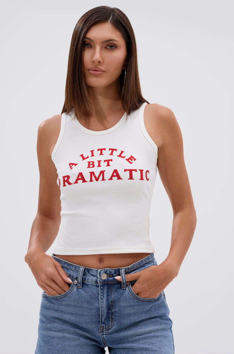 A woman with straight brown hair wears the A LITTLE DRAMATIC EMBELLISHED TANK TOP, featuring a white lace-up back and bold red lettering, paired with blue jeans and standing casually against a plain background. 0