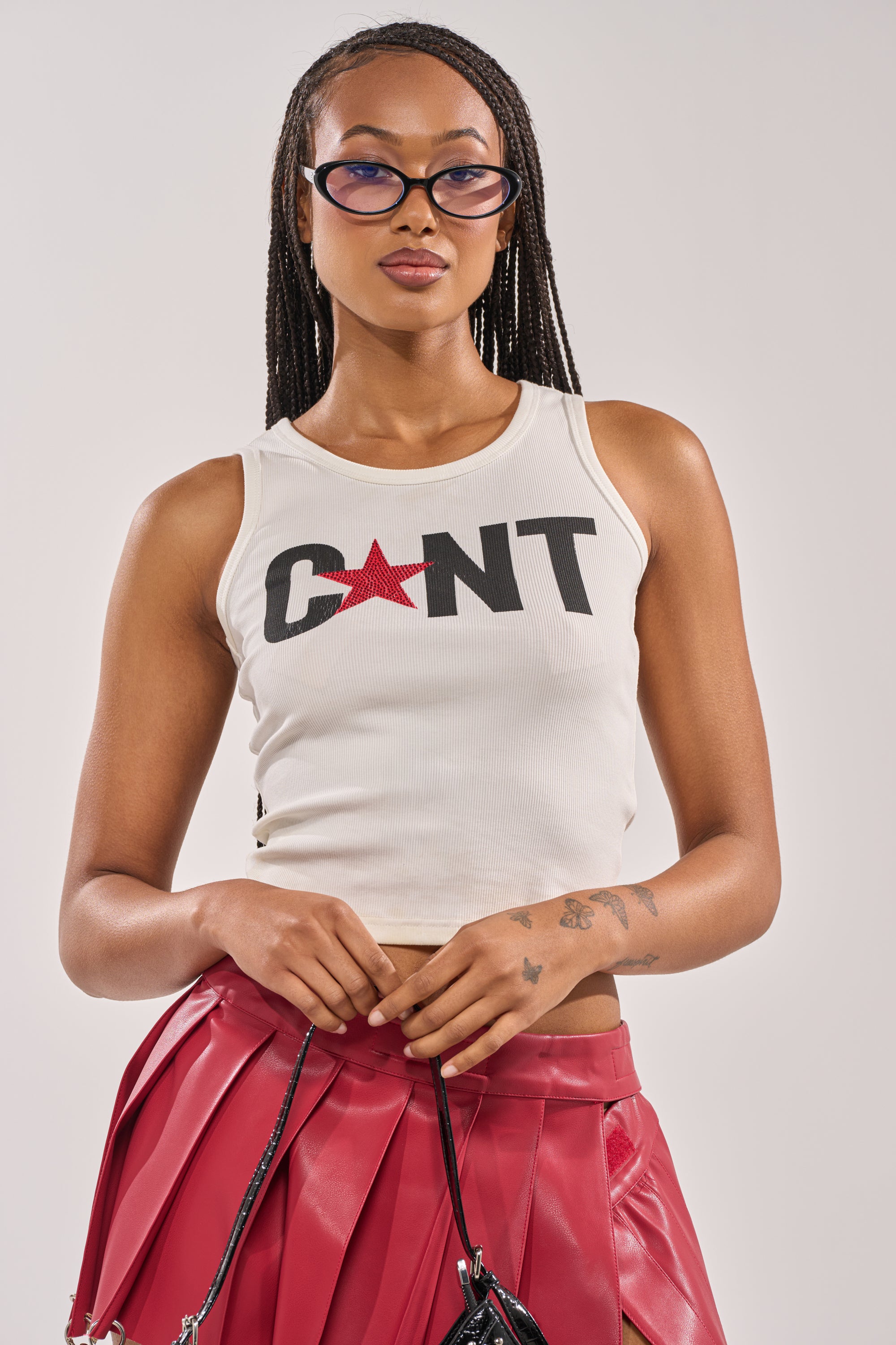 A woman with braided hair wears cat-eye sunglasses, the CNTY LACE UP TANK featuring bold black “C*NT” text and a red star, plus a pleated red skirt, standing against a plain background. 2