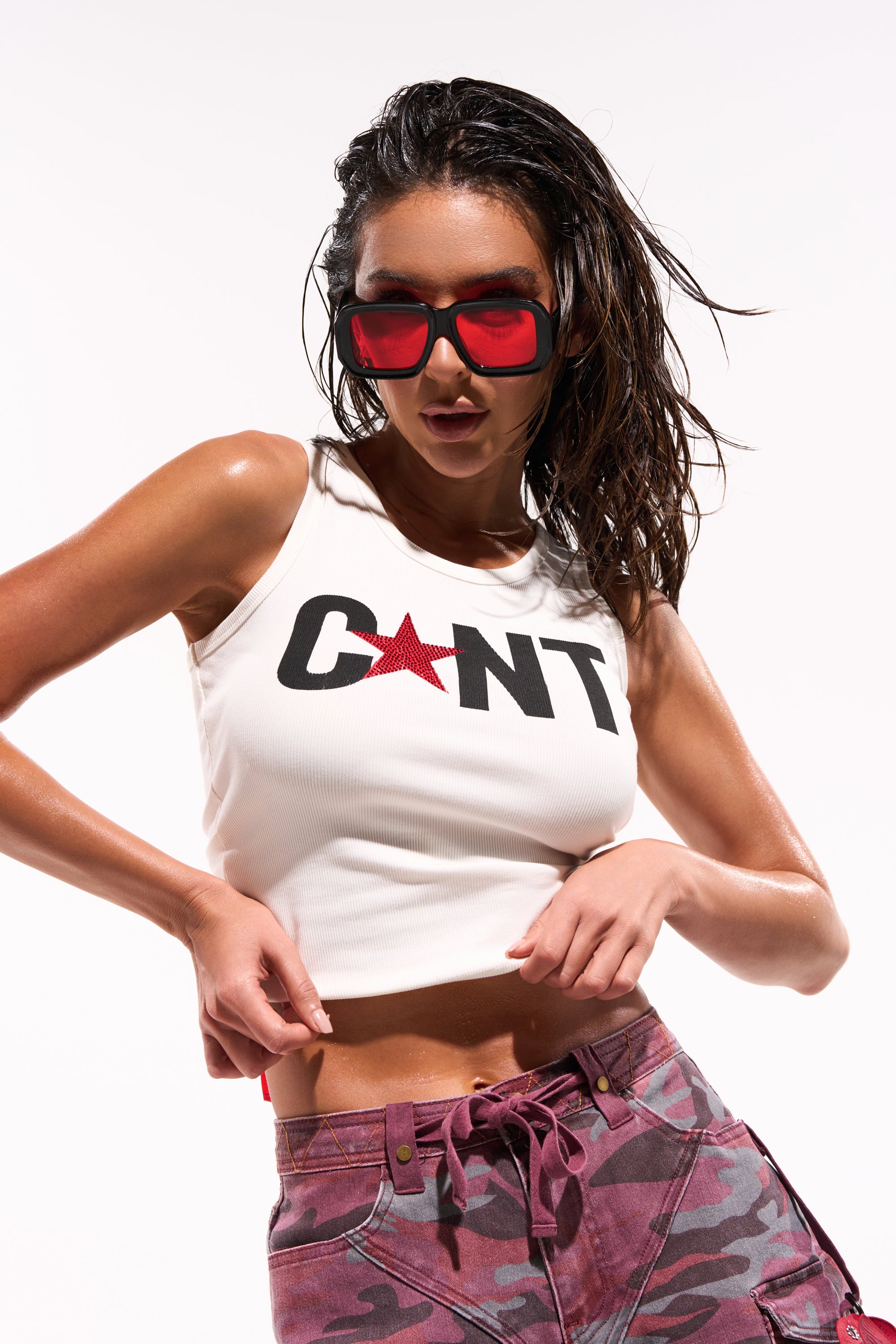 A woman with wet hair and large red sunglasses poses against a bright white background, wearing the CNTY LACE UP TANK with a red star and pink camo pants. 0