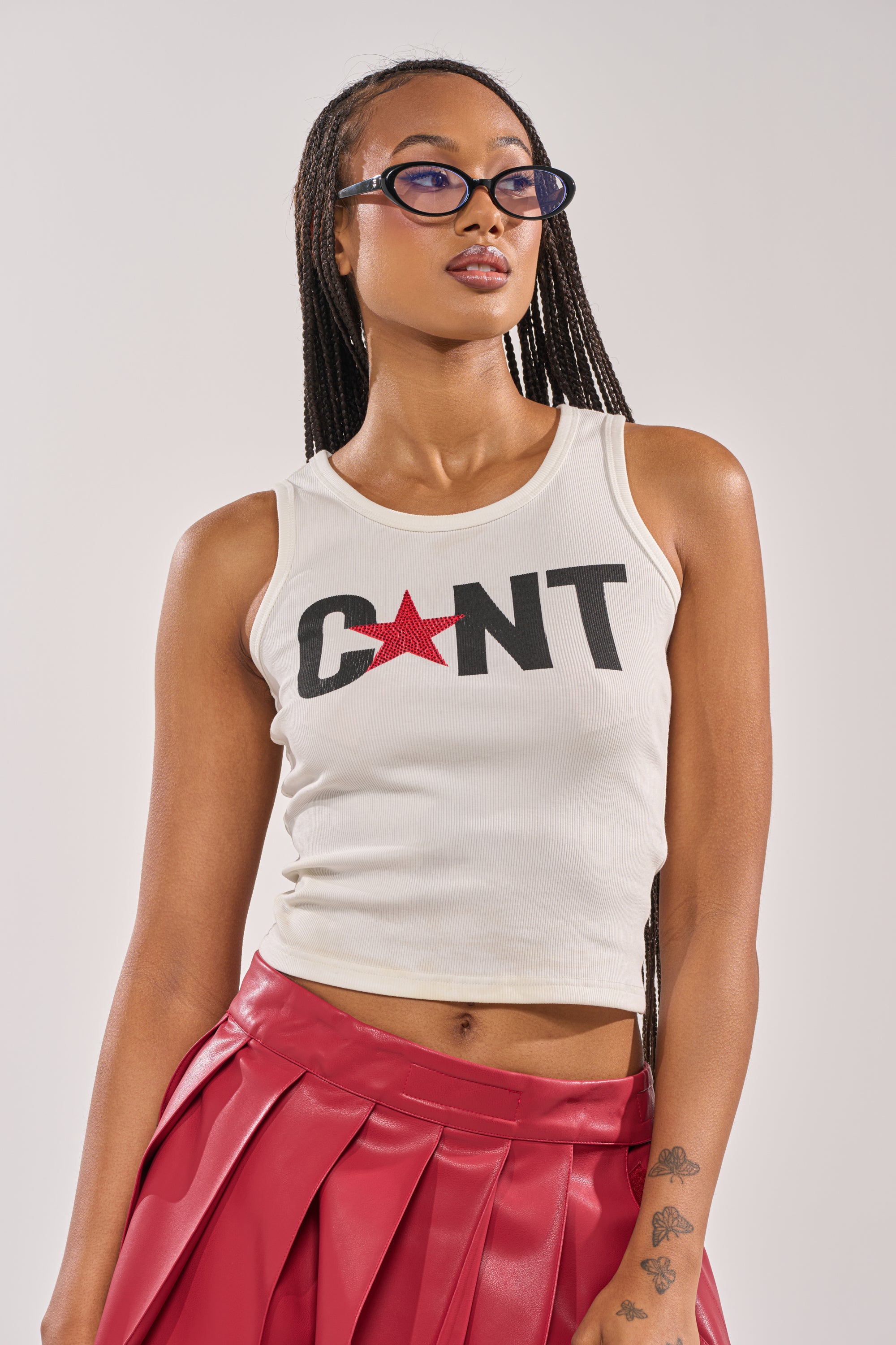 A woman with long braids and glasses wears the CNTY LACE UP TANK, featuring "C*NT" with a red star for the “U,” paired with a red pleated skirt. She has tattoos on her left forearm and gazes off to the side. 3