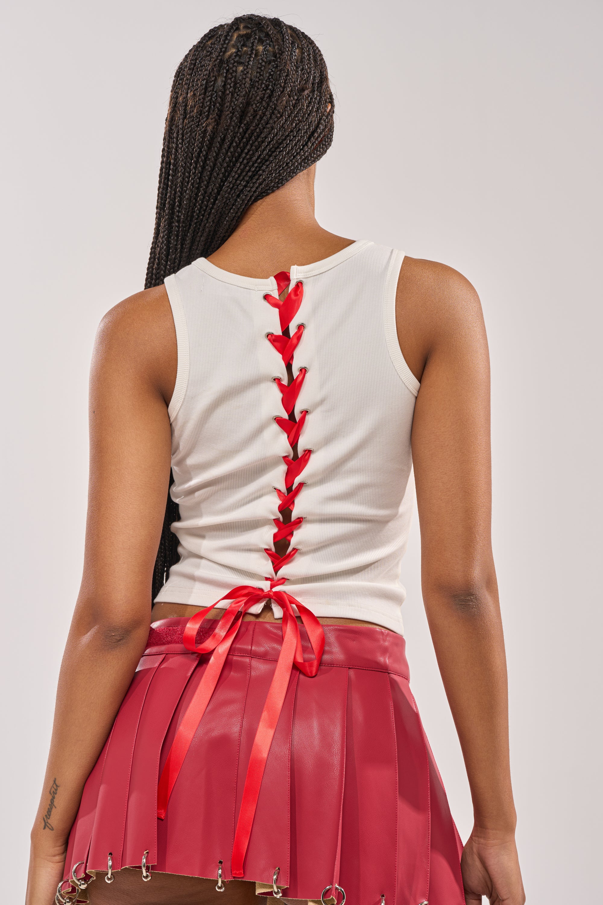 A woman with braided hair is seen from the back, wearing the CNTY LACE UP TANK—a sleeveless white top laced with red ribbon down the center—paired with a red pleated skirt featuring silver ring details on the hem. 5