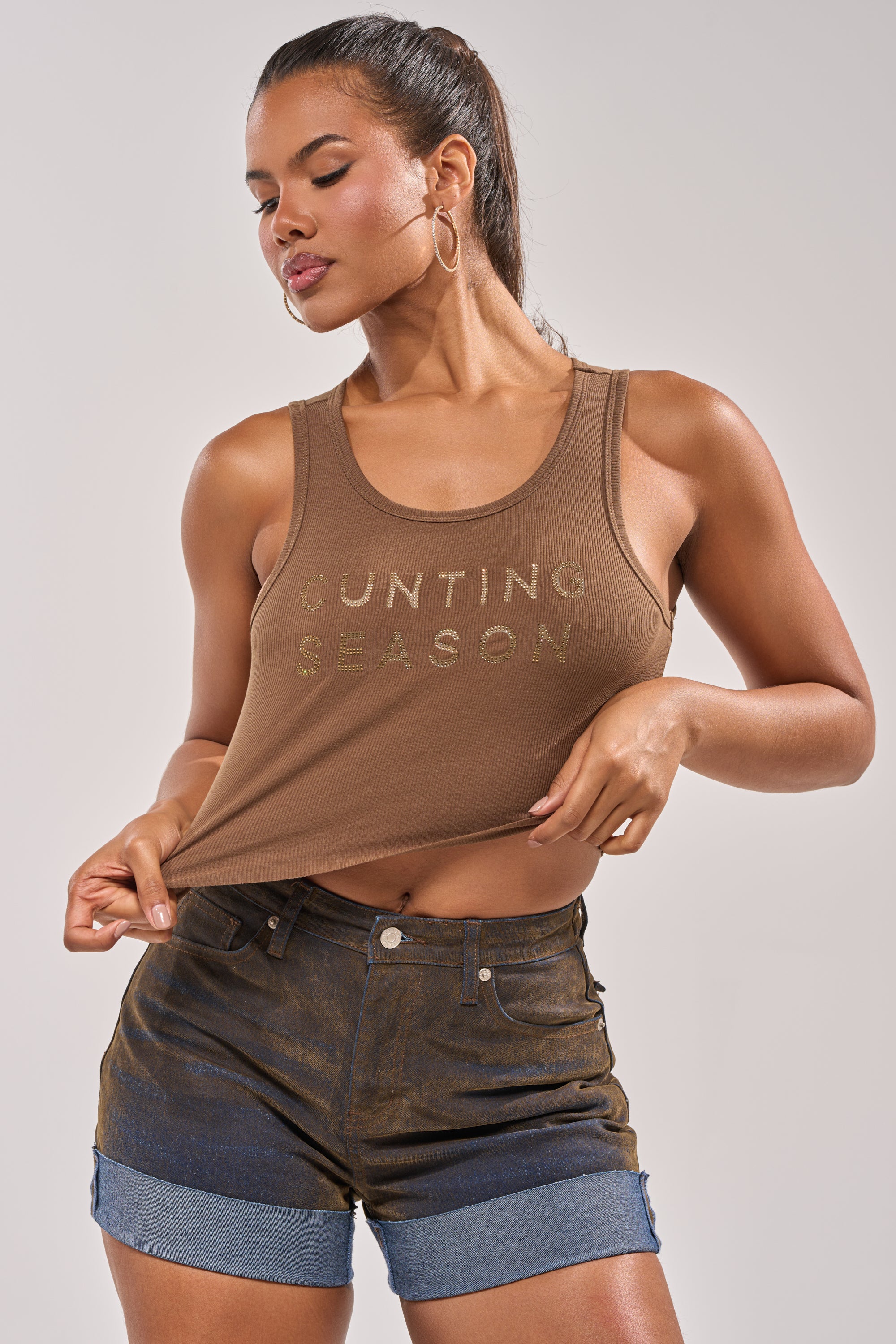 A woman models the CNTING SZN RHINESTONE RIBBED TANK in brown with dark denim shorts, holding the hem and looking to the side. She completes her look with hoop earrings. 1