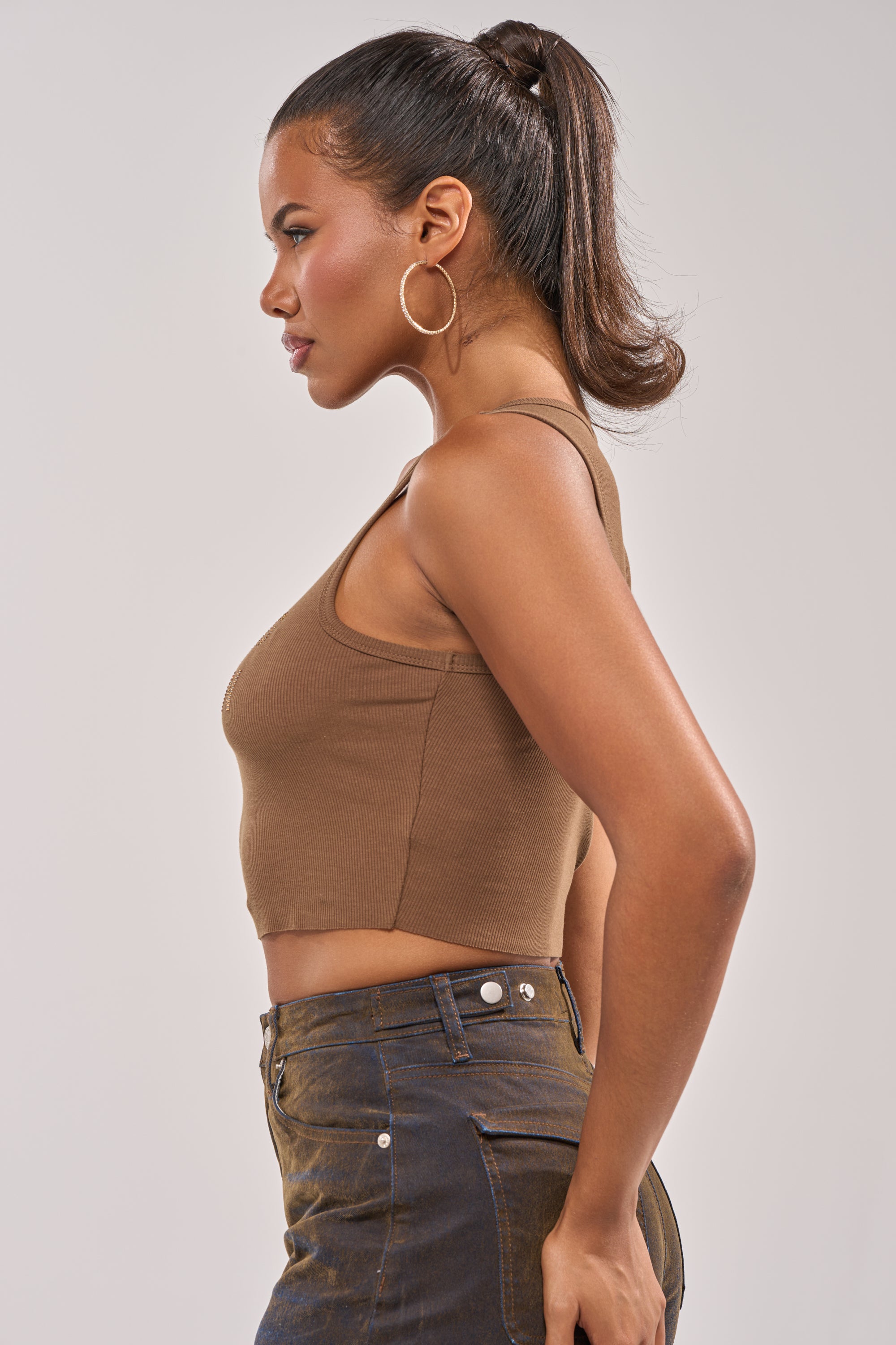 Woman in profile wearing the CNTING SZN RHINESTONE RIBBED TANK, high-waisted dark jeans, large hoop earrings, and a high ponytail, posed against a plain light background. 4