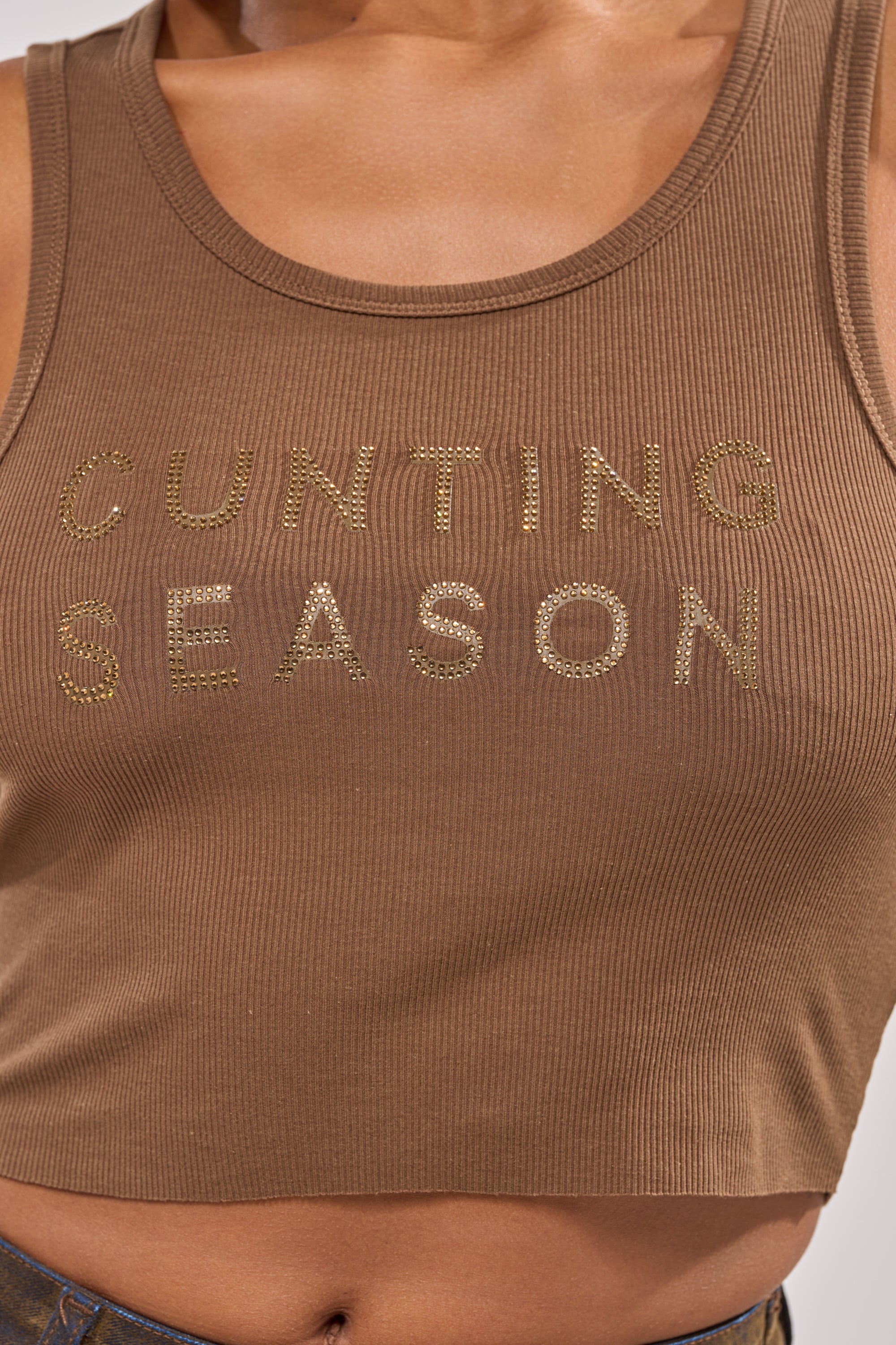 A close-up of a person wearing the CNTING SZN RHINESTONE RIBBED TANK, a brown ribbed crop top featuring gold rhinestones spelling out "CNTING SZN" across the chest. 8