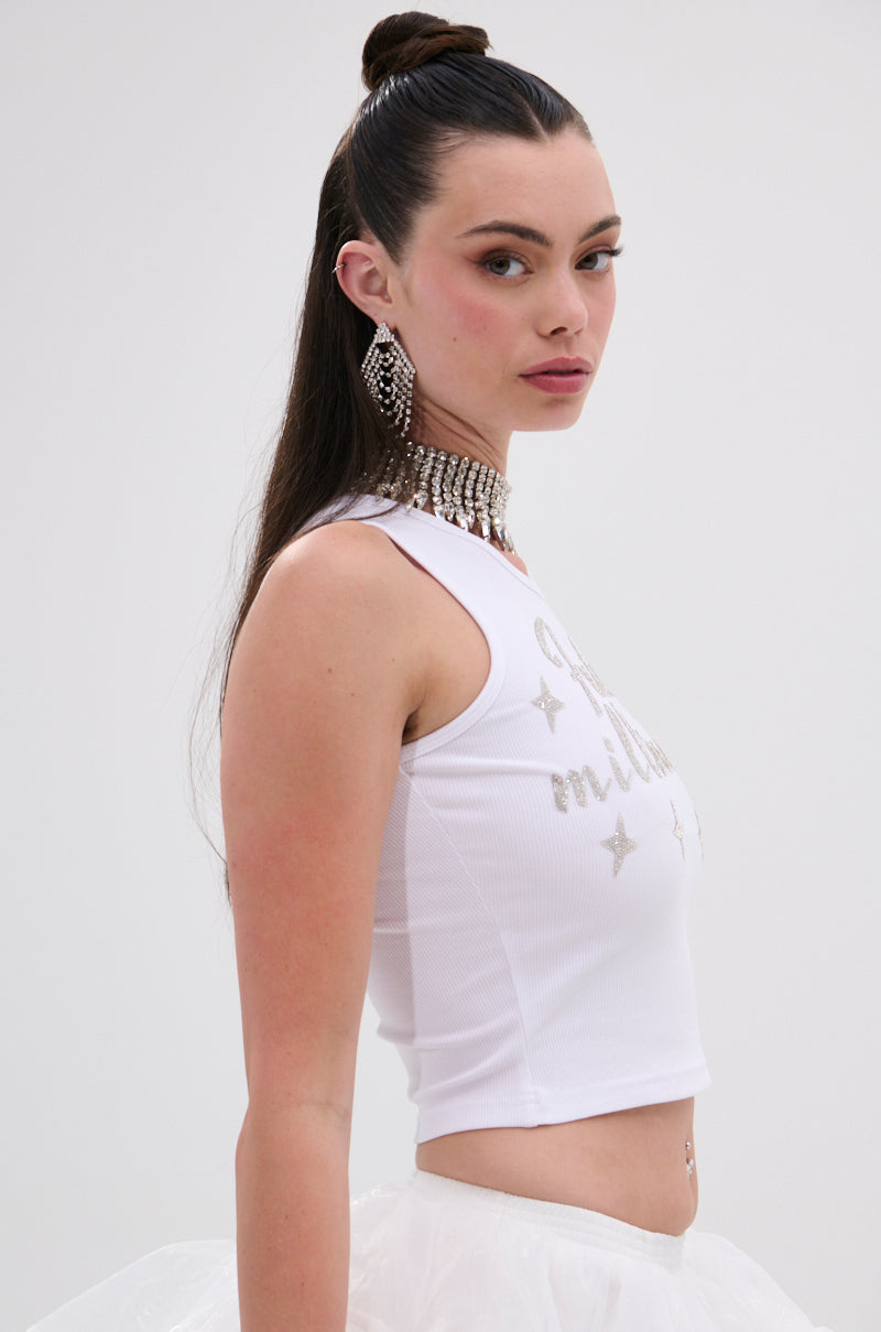 A young woman with long dark hair in a high bun poses sideways against a plain background, looking at the camera. She wears rhinestone earrings, a white skirt, and the FUTURE MILLIONAIRE TANK TOP—radiating future millionaire vibes. 2