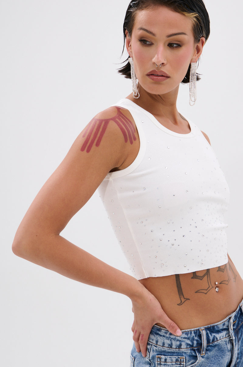 A person with short dark hair wears the RHINESTONE QUEEN TANK and blue jeans, showcasing arm and torso tattoos. Sporting long earrings and a confident pose against a light background, they channel bold Y2K fashion vibes. 2