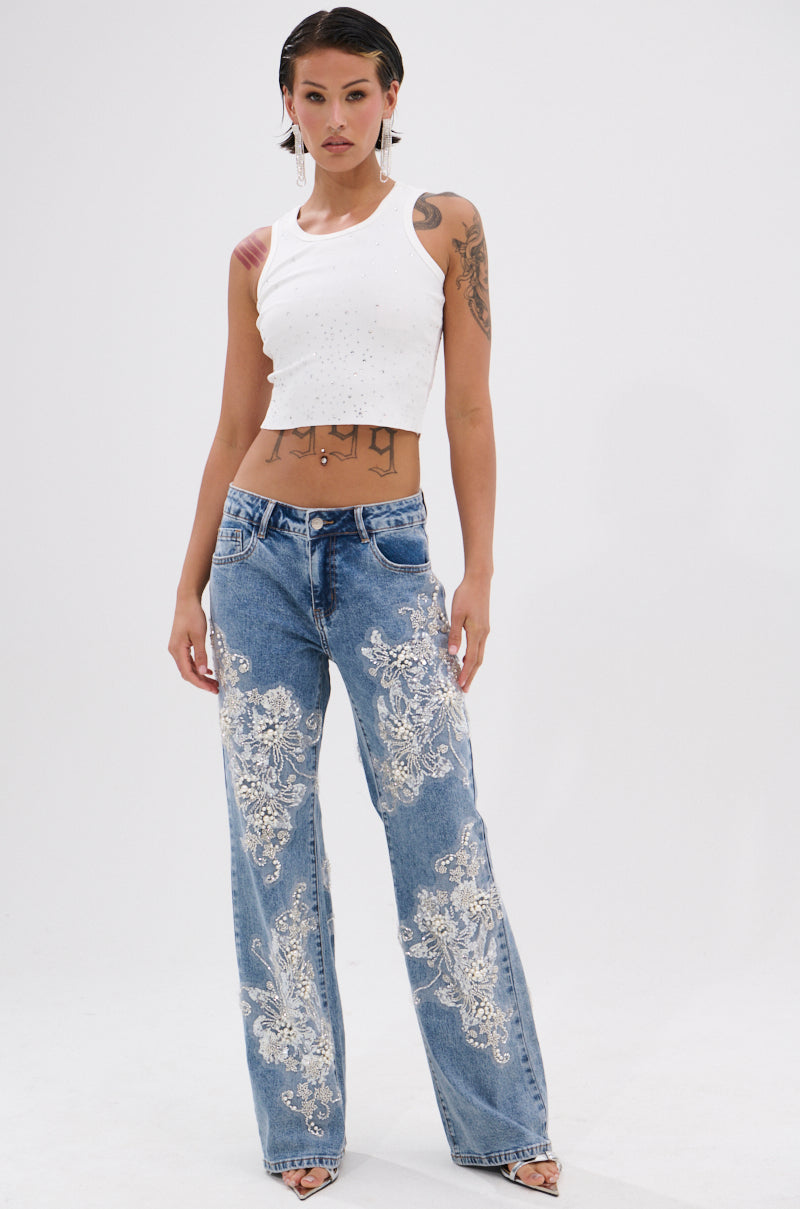 A woman with short dark hair and visible tattoos wears the RHINESTONE QUEEN TANK, a Y2K-style white sleeveless cropped top, paired with high-waisted blue jeans featuring intricate white floral embroidery. She stands against a plain white background. 4
