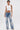 A woman with short dark hair and visible tattoos wears the RHINESTONE QUEEN TANK, a Y2K-style white sleeveless cropped top, paired with high-waisted blue jeans featuring intricate white floral embroidery. She stands against a plain white background.