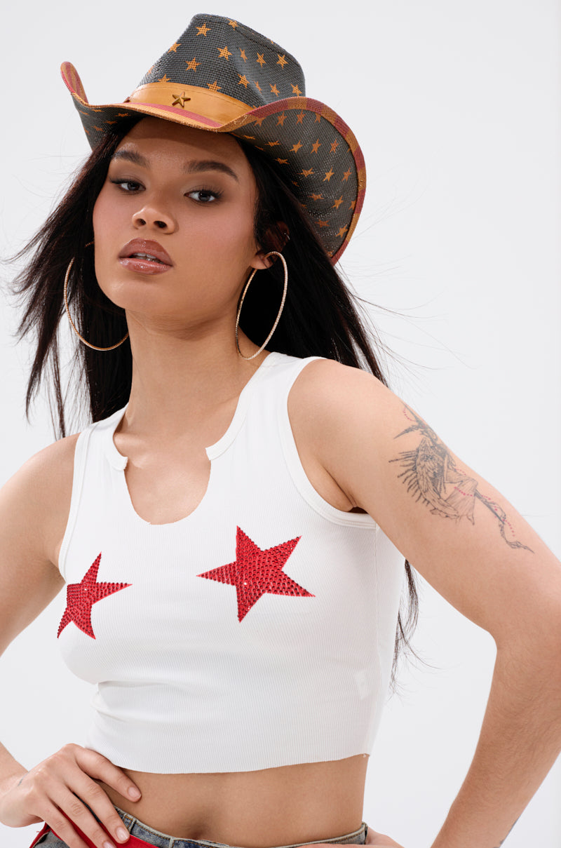 A woman with long dark hair, a tattooed upper arm, and large hoop earrings wears the SEEING STARS CROPPED TANK TOP—a white top featuring two red glitter star appliqués—styled with a star-patterned cowboy hat for a bold Western-inspired look.